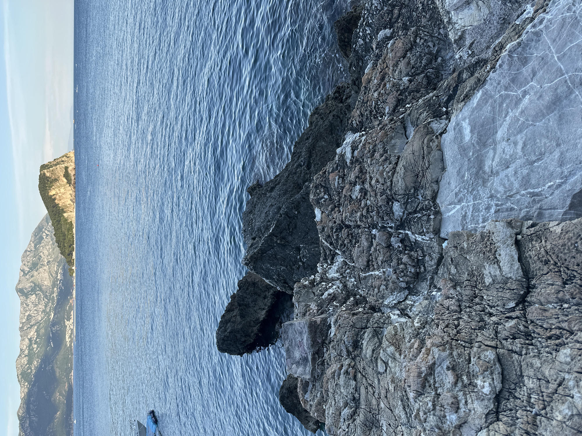 The image depicts a rocky coastline in Budva, Montenegro, viewed from an elevated perspective. A portion of a mountain range is visible in the background under a light blue sky, separated from the rocks by water. The foreground features a jagged rock formation with various textures and colors, ranging from dark gray to lighter shades. The water is a deep blue with subtle ripples. A small boat or vessel can be seen in the distance along the coastline near the mountains.