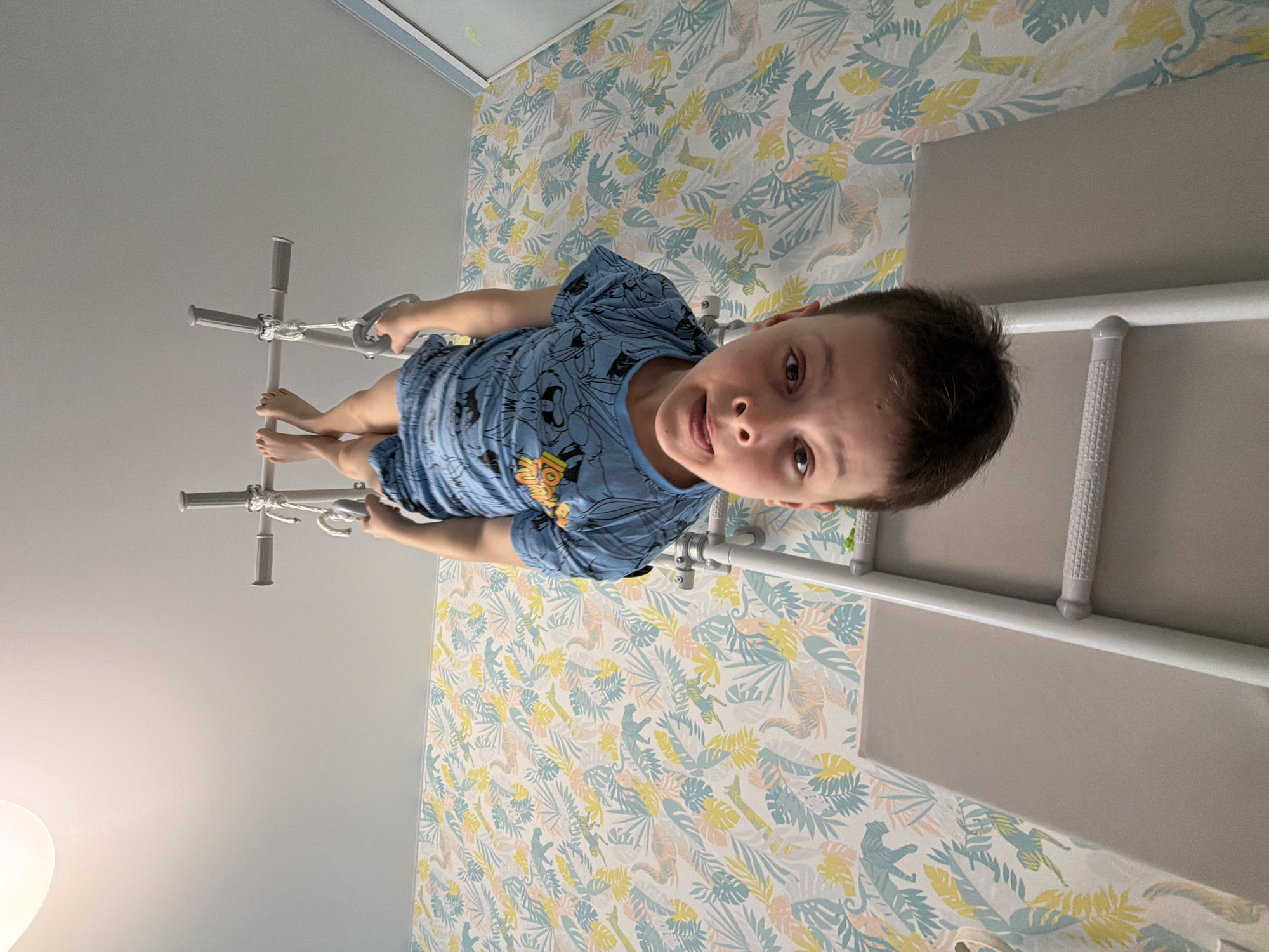 A child is hanging from a white physical therapy ladder in a room with wallpaper featuring a leafy pattern in blue, yellow, and pink. The ladder is attached to a gray padded bench. The child is wearing a blue t-shirt with cartoon characters. The location of the scene is in Votkinsk, Russia.
