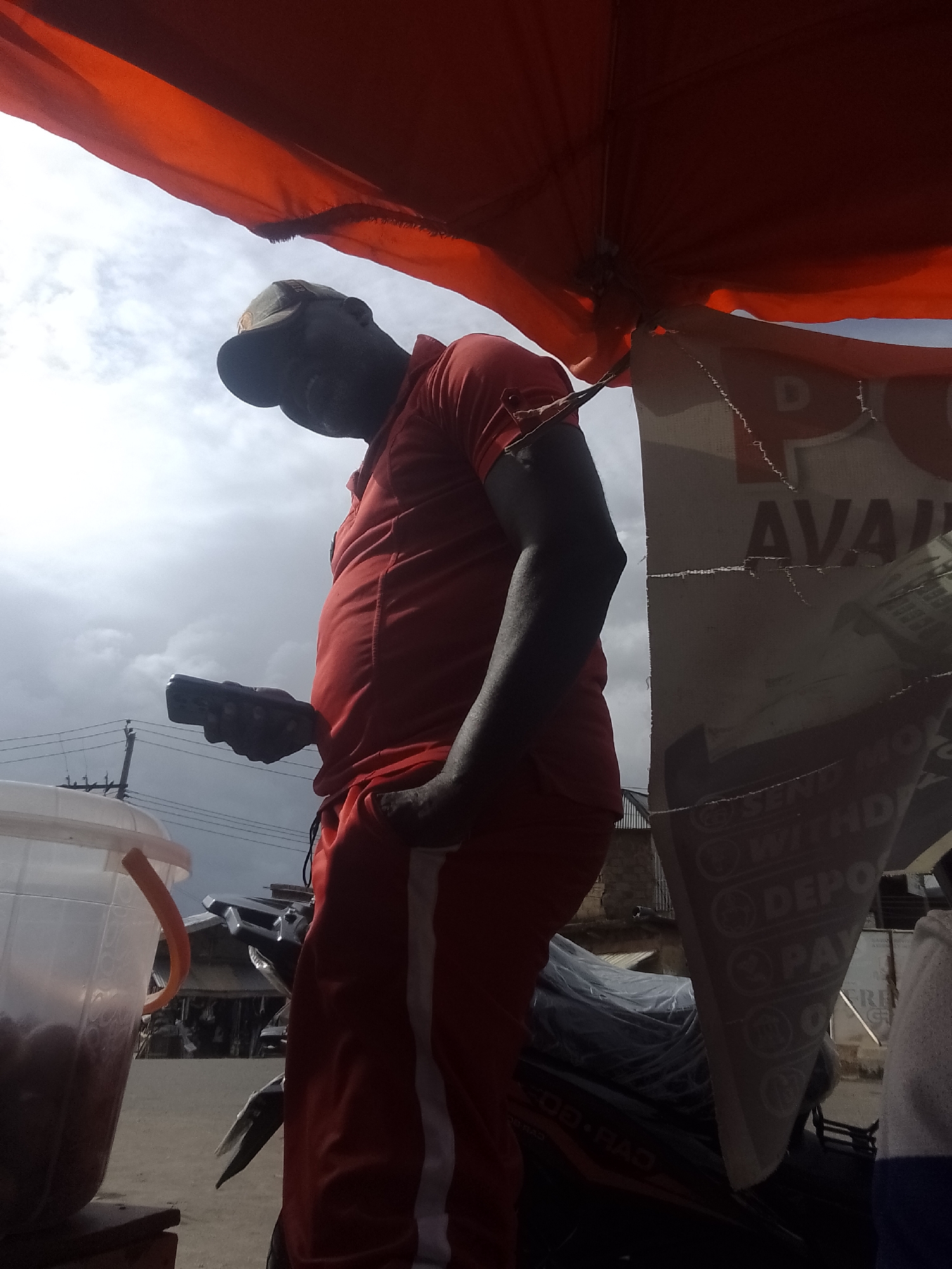 An outdoor, medium shot shows a dark-skinned man in a red shirt and red pants with white stripes, wearing a dark baseball cap, holding a black smartphone in his right hand. He is standing under a large, orange canopy that casts a shadow over the scene. The man is looking down at the phone, and his left hand is in his pants pocket. To the left of the man is a large, clear plastic bucket filled with a dark red liquid, with an orange handle. Behind the man, a motorcycle is partially visible, covered with a plastic sheet. To the right of the man, a torn, white poster with black lettering is affixed to a structure, with partial words like "AVA" and "WITHD" visible. The background shows a cloudy sky, power lines, and some buildings. The scene appears to be in a street market or an outdoor vendor stall.