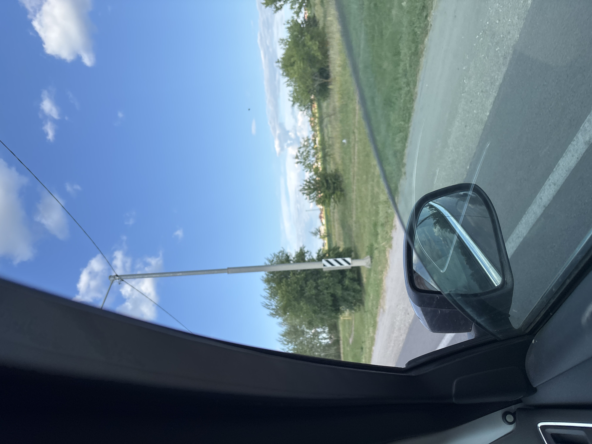 The image captures a view from inside a car traveling down a road. The scene is set outdoors under a bright blue sky with scattered white clouds, suggesting a clear day. The time of day appears to be daytime, likely late morning or early afternoon, given the strong sunlight.
The prominent subjects include the car's side mirror, reflecting the sky and trees, and the road with a white lane marking. Beyond the road, there is a grassy area with several trees and a tall pole with a black and white striped marker. The landscape suggests a rural or semi-rural setting. The overall mood is calm and ordinary, depicting a typical journey.
The image is oriented vertically. Visible text is not present in the image. The location context is Novaya Usman, Russia.
