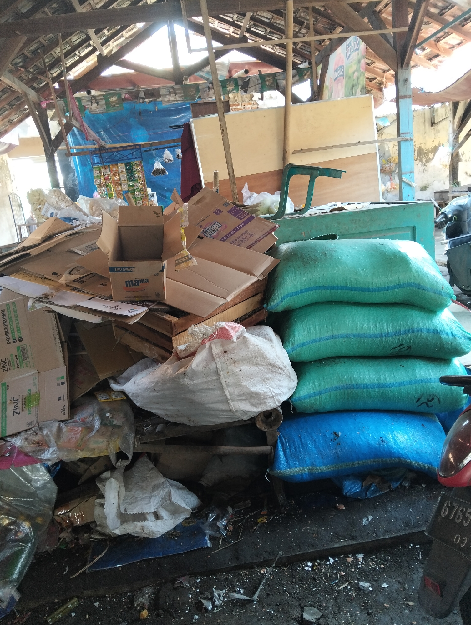 The image depicts a cluttered, open-air storage area, likely an outdoor market or warehouse in Muncar, Indonesia, characterized by a dilapidated wooden roof structure with visible beams and corrugated metal sheeting. Sunlight streams through gaps in the roofing, illuminating piles of cardboard boxes, plastic packaging, and several large, stacked sacks filled with goods. The sacks are primarily teal and blue, with blue stripes. A red scooter is partially visible in the lower right corner, displaying a license plate with the number "6765 09". The ground is a mixture of dirt and debris, with scattered smaller items like broken shells or food scraps. Various cardboard boxes are piled high, some open, revealing their contents or previous use. One box prominently displays the brand name "mama". A blue tarp is visible in the background, along with some hanging items that appear to be small packages or snacks. The overall scene suggests a disorganized storage or distribution point for goods within a potentially commercial or industrial setting in Muncar.