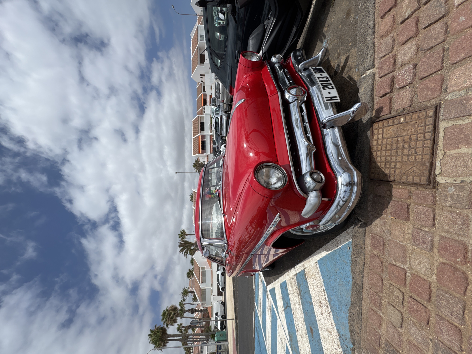 A red 1950s-era Ford Fairlane is parked in a street lot in Arona, Spain, with white buildings and palm trees in the background. The car is facing forward, with its chrome bumper and headlights prominently displayed. The parking space is marked with blue and white stripes. Other cars are visible in the background, along with a dark-colored scooter parked next to the Ford. The sky is blue with white, fluffy clouds.