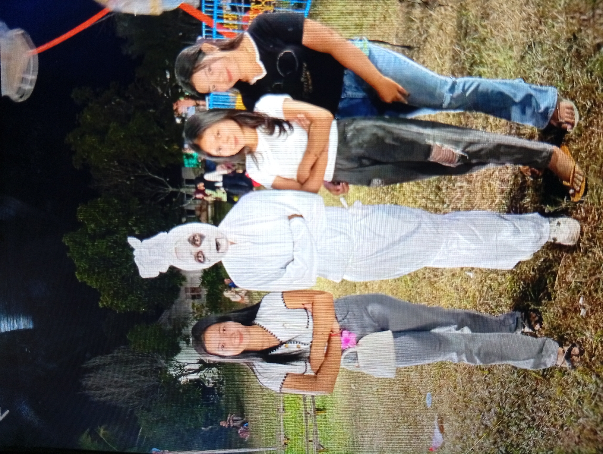 In Lhokseumawe, Indonesia, four individuals are depicted outdoors on a grassy area, possibly a park or open field. Two young women are standing, facing forward and smiling, with one wearing a black t-shirt and jeans, and the other in a white t-shirt and dark pants. Behind them, two other individuals are posed lying down. One is dressed in a white costume, resembling a ghost or mummy, with visible eyes and mouth. The second person lying down is a young woman wearing a striped shirt and grey pants, holding a pink flower and a white bag. The background includes trees and some structures, suggesting an outdoor event or gathering. The lighting indicates it is daytime, with sunlight illuminating the scene.