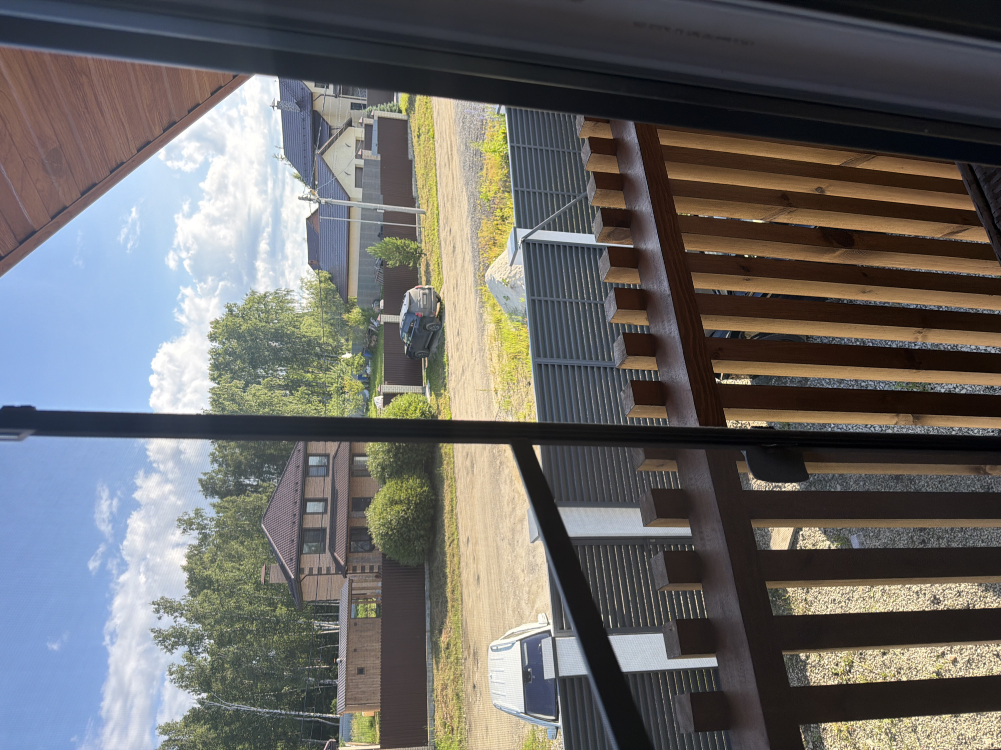 The image depicts a view from a wooden balcony overlooking a suburban street in Malinovka, Russia. The balcony features a wooden railing with square posts. Beyond the balcony, a grey corrugated metal fence separates the property from the street. A white vehicle is parked on the street in front of a brown fence and a small wooden outbuilding. Further down the street, a dark-colored SUV is parked near a row of houses with brown roofs. Lush green trees line the background, and the sky is blue with scattered white clouds. The scene appears to be set in the daytime under sunny conditions.