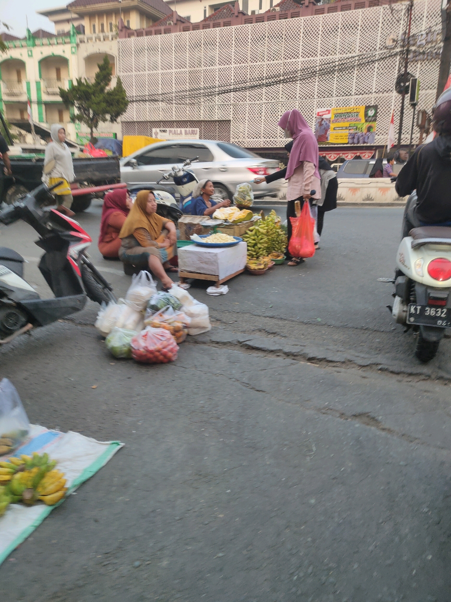 In Samarinda, Indonesia, a street scene unfolds with an informal market. A woman wearing a pink hijab and a light-colored top stands near a fruit stall, interacting with another person. The stall displays a variety of fruits, including bananas and what appear to be durians, arranged on a white surface and in bowls. Nearby, three women are seated on the ground, some with food items. One woman, in an orange hijab, is holding a plate. Another woman, in a red hijab, is looking towards the fruit stall. A pile of plastic bags filled with produce, including tomatoes and green vegetables, sits on the asphalt. Two motorcycles are present; one is parked and partially visible in the foreground, while another, a white scooter with the license plate "KT 3632," is positioned further back. The background shows a building with white lattice-like detailing, typical of Indonesian architecture, and a street with visible cracks. A silver car is also parked on the street. The overall atmosphere suggests a bustling local market scene.