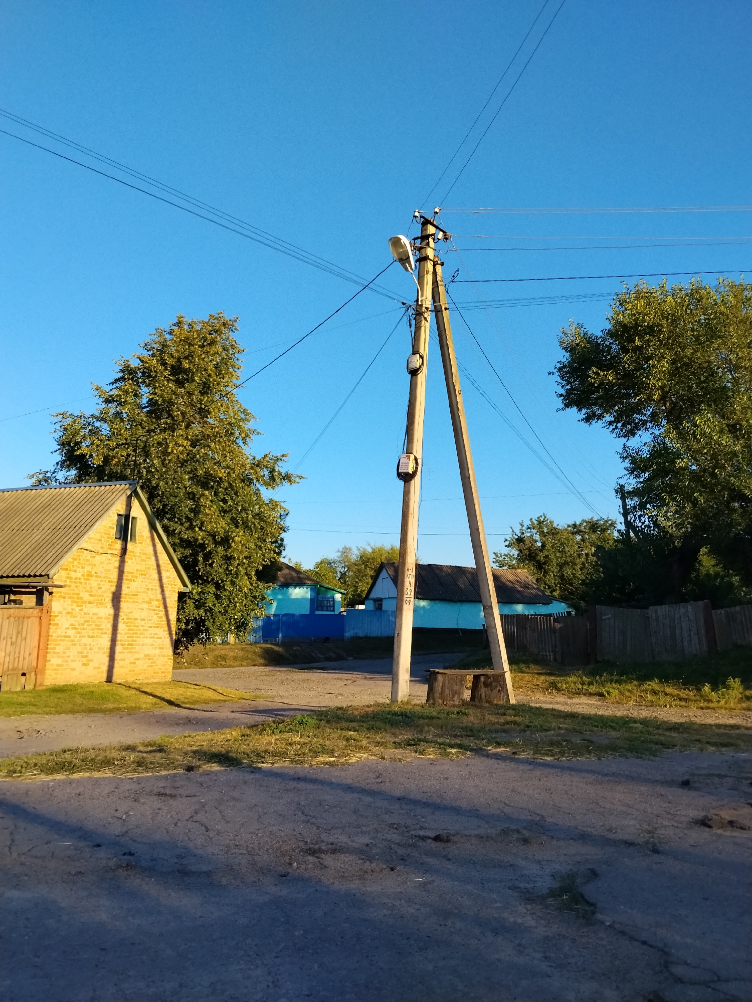 The image depicts a street scene in Ustyvytsia, Ukraine, under a clear blue sky. A wooden utility pole, triangular in structure, stands prominently in the foreground, supporting electrical wires and two utility meters. A streetlamp is attached to the top of the pole. To the left of the pole, a yellow brick building with a gray corrugated roof is visible, with a wooden fence in front of it. Behind the brick building, a large, leafy tree dominates the mid-ground. Further back, a bright blue fence runs horizontally, separating the foreground from several houses with dark roofs. These houses appear to be single-story structures. The ground in the foreground is a dirt road with patches of grass and dried vegetation. Shadows are cast across the road, indicating the direction of sunlight. The overall impression is of a rural or suburban street in a village.