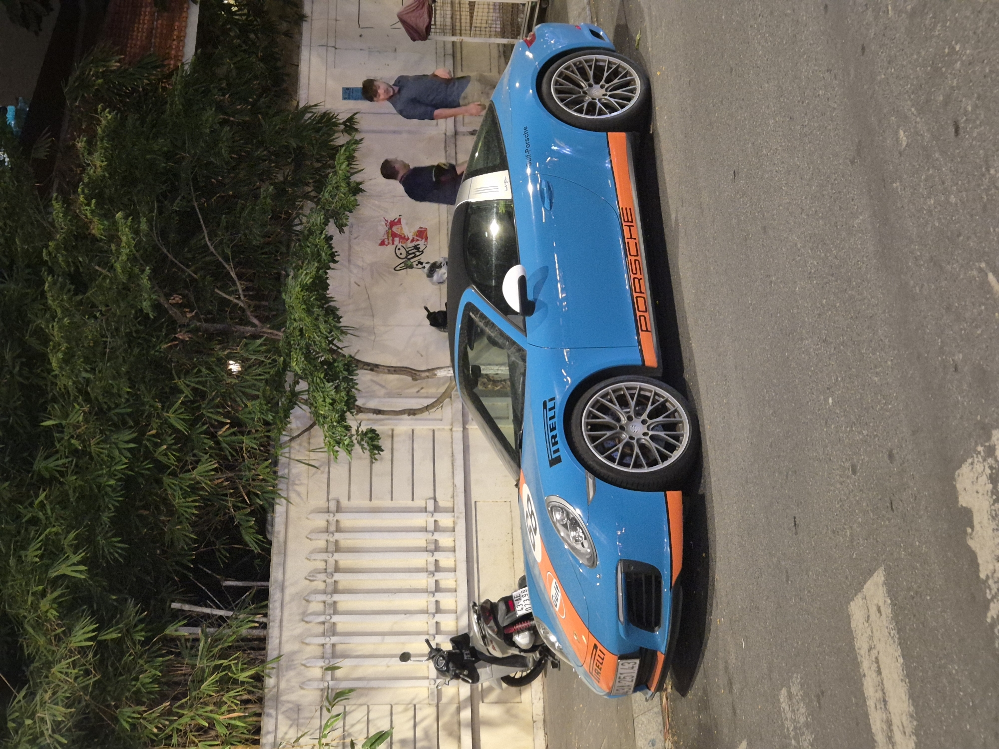 The image shows a blue Porsche 911 Targa 4 GTS with orange accents, parked on a street in Đà Nẵng, Vietnam, at night. The car has racing decals, including "Pirelli" and "Gulf-Porsche" branding, and a number "88" on the hood. A white scooter is parked behind the Porsche. Two men are standing near a white fence in the background, and a tree with dense foliage is visible above the fence. The street appears to be a pedestrian crossing. The lighting suggests it is evening or night, with artificial lights illuminating the scene.