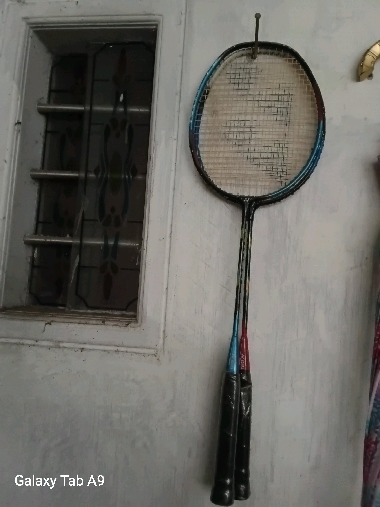 A badminton racket with blue and black accents and a Yonex logo is hanging on a nail on a textured, light-colored wall. The racket strings appear worn. To the left of the racket is a window with metal bars and a decorative glass pane. The bottom left corner of the image displays the text "Galaxy Tab A9".