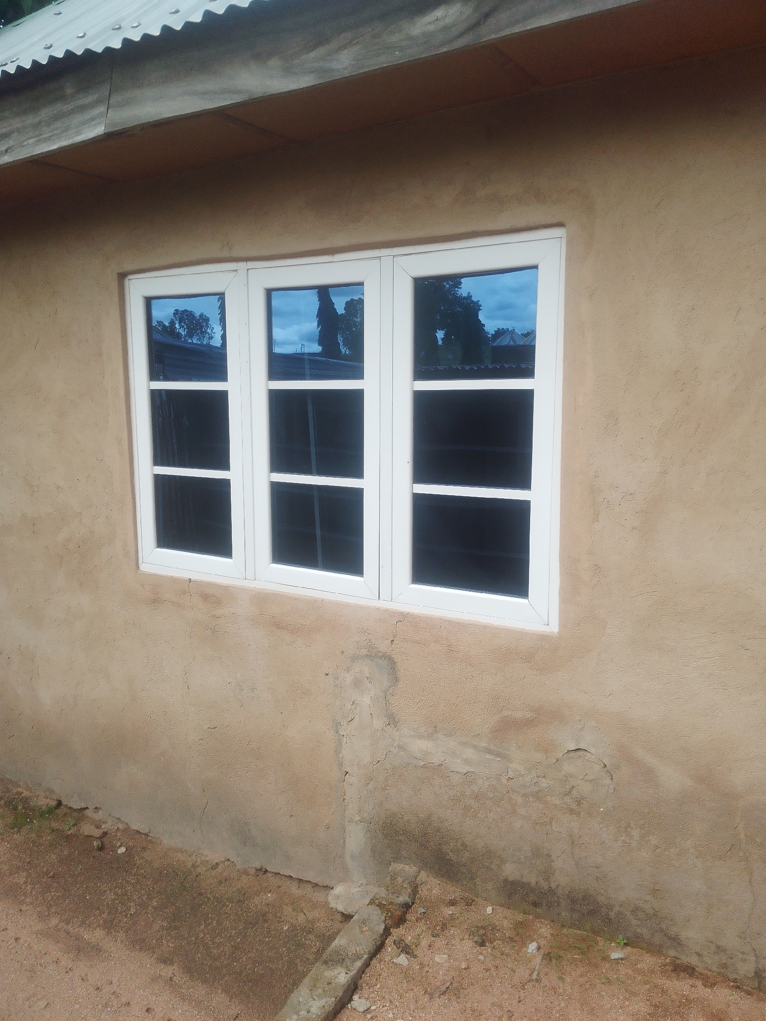 A three-panel window with white frames is installed in the tan-colored, rough-textured wall of a building. The glass in the windows is tinted blue and reflects the sky and trees. The building's roof, made of corrugated metal, is visible above the window. The ground in front of the building is a mixture of dirt and small stones. The scene appears to be outdoors in daylight, with the weather likely clear as indicated by the sky reflection. The location is Jalingo, Nigeria. There is no visible text in the image.