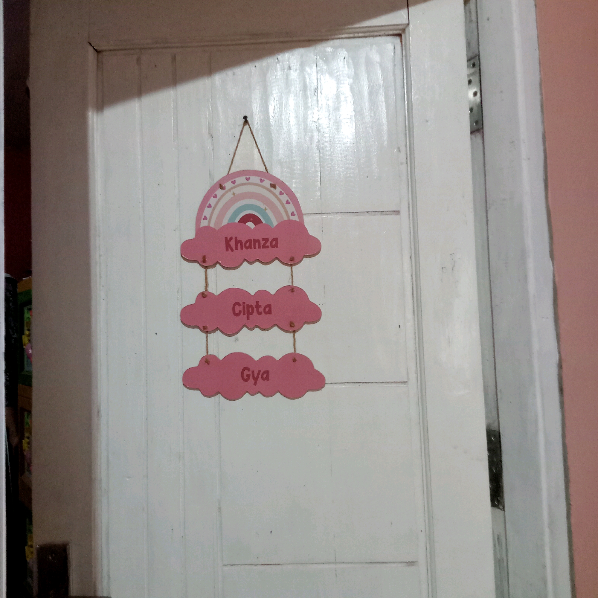 The image shows a decorative sign hanging on a white wooden door. The sign is shaped like a rainbow with clouds below it. Each cloud has a name written on it in pink lettering. From top to bottom, the names are "Kanza", "Cipta", and "Gya". The sign is hung by twine from a small nail. The door appears to be in a residential setting, possibly a bedroom, in Purwakarta, Indonesia, as indicated by the provided location. The lighting suggests an indoor environment, with a shadow cast from the left side of the frame onto the door. There are no people visible in the image.