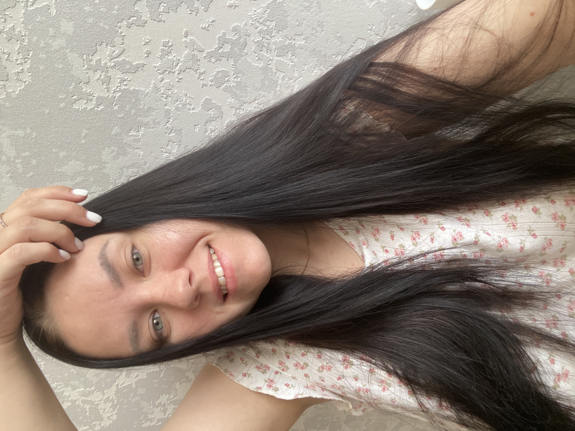 A woman with long, dark hair is smiling at the camera. She is wearing a white top with a small floral print. Her left hand is near her forehead, and she has white nail polish. The background is a textured, light gray wall. It is not possible to confirm the location in the city of Chaikovsky, Russia based on this image alone.