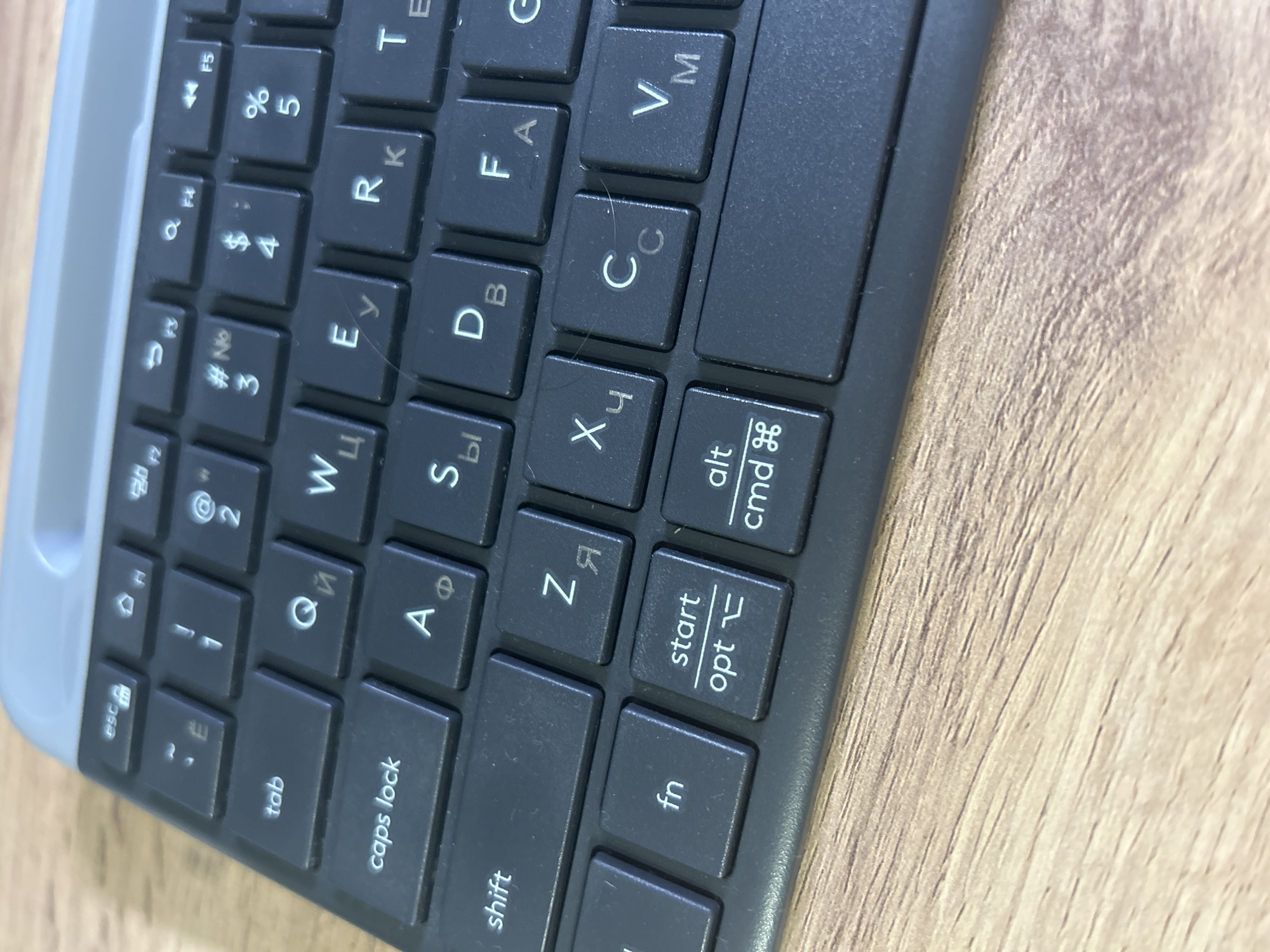 The image shows a close-up, slightly angled view of a dark gray computer keyboard, likely a wireless or compact model, resting on a light brown wooden surface. The keyboard has a top edge that is a lighter gray or silver color. The keys are visible, displaying a mix of Latin and Cyrillic characters, along with standard function and modifier keys. Specifically, prominent keys include "esc," "tab," "caps lock," "shift," "fn," "start/opt," and "alt/cmd." Individual letter keys such as "Q," "W," "E," "R," "T," "A," "S," "D," "F," "G," "Z," "X," "C," "V," "M," and several others are identifiable. Some keys also show symbols and numbers like "1," "2," "3," "4," "5," "%," and "#." The lighting suggests an indoor environment. There are no people, animals, or other significant objects visible in the frame. The wooden surface appears to be a tabletop or desk. The overall impression is one of a functional workspace.