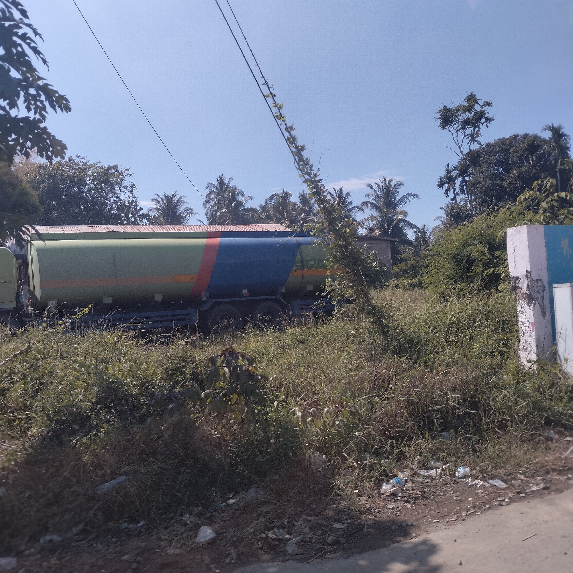 The image presents an outdoor scene in Cotpuuk, Indonesia, under a clear, bright blue sky on a sunny day. The central subject is a large tanker truck, primarily painted green, featuring prominent red and blue vertical stripes on its side, with a narrow orange stripe also visible. The truck is situated in an overgrown area dominated by tall grass and weeds, obscuring its lower half.
In the background, a dense canopy of tropical trees, including numerous palm trees, extends across the horizon, suggesting a lush, verdant environment. Overhead, thin black power lines traverse the sky, with noticeable vines growing and clinging to one of them, indicating natural overgrowth or a lack of maintenance.
To the far right, a sturdy concrete pillar, painted white with remnants of blue paint on one side, stands partially damaged and weathered. The foreground reveals a dirt or gravel path/roadside that leads into the scene, where scattered pieces of litter, such as plastic bottles and bags, are visible among the sparse vegetation and on the ground. The strong sunlight creates clear shadows. No people or discernible text are visible in the photograph. The overall impression is a quiet, somewhat rural or semi-rural setting experiencing natural encroachment and some neglect.