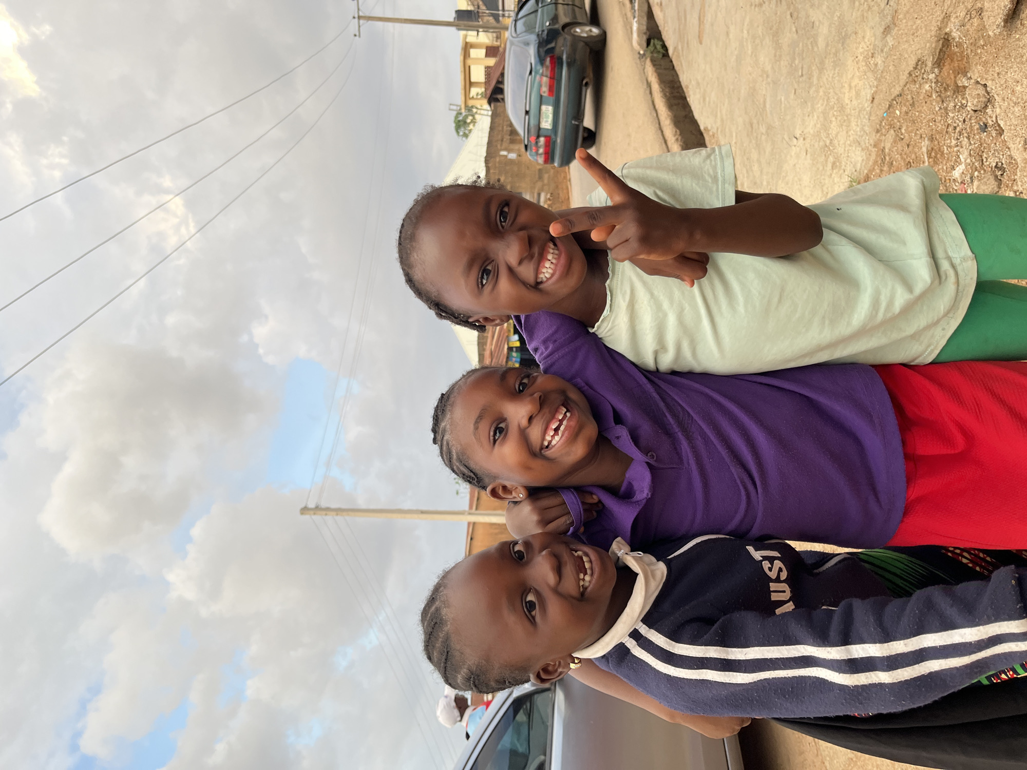 The image, rotated 90 degrees counter-clockwise, depicts three young girls happily posing for a photograph on a street in Jos, Nigeria. All three girls are smiling brightly at the camera, conveying a sense of joy and playfulness.
The girl positioned at the top of the rotated frame wears a light green t-shirt and green bottoms, making a peace sign with her right hand. The girl in the middle is dressed in a purple polo shirt and red bottoms, with her arm around the youngest girl. The girl at the bottom of the frame wears a navy blue long-sleeved shirt with white stripes on the sleeves and a white collar. The word "AUST" is visible in white capital letters on her shirt.
The background shows a street scene with a dark-colored sedan parked to the upper right. The car's license plate appears to read KJJ-273XX. Behind the car, buildings are visible, suggesting an urban or semi-urban environment. Power lines extend across the upper left portion of the frame, set against a cloudy sky with hints of blue. The lighting suggests it is daytime, with a soft, diffused light.