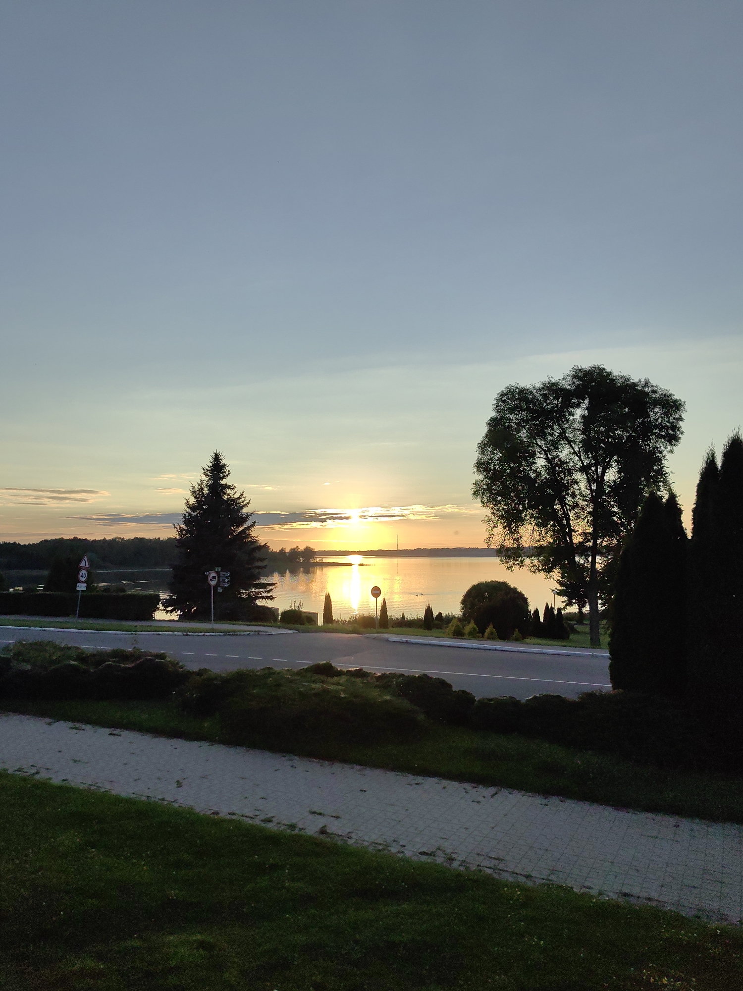 This is a landscape photograph of a sunset over a body of water, likely a lake, in the city of Myadzyel, Belarus. The sun is low on the horizon, casting a warm, golden glow across the water and sky. A silhouette of trees and foliage lines the distant shore. In the foreground, a paved walkway runs alongside a road. There are several traffic signs and a large conifer tree along the roadside. The overall scene is serene and picturesque, typical of a lakeside setting at dusk.