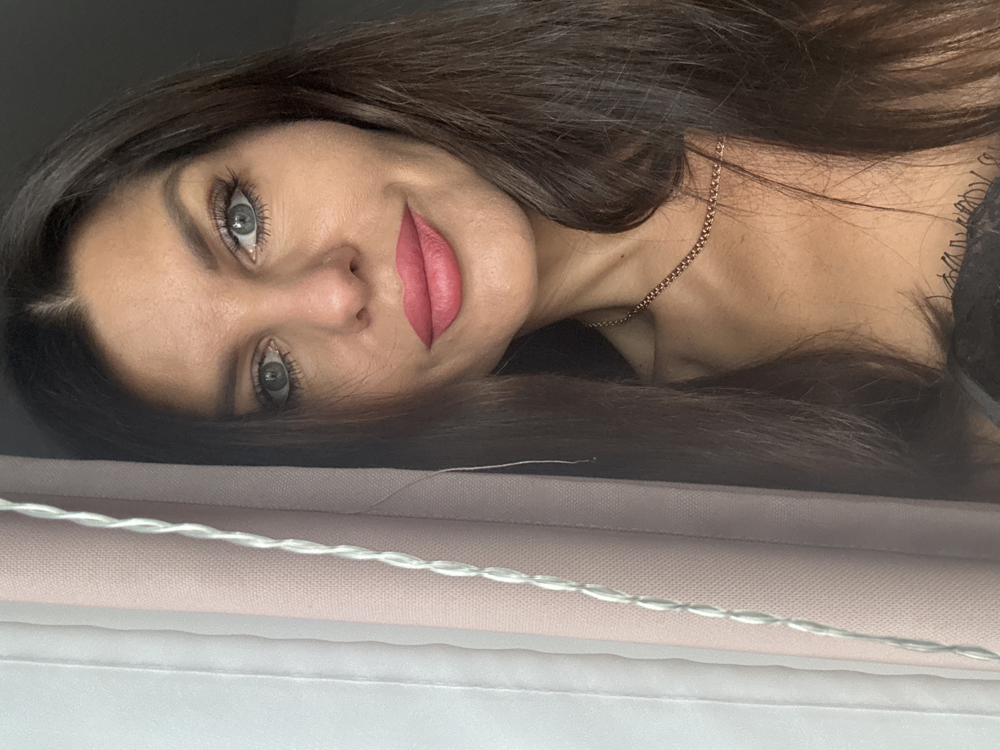 A medium shot image shows a woman with long, dark hair and striking blue eyes. She is looking directly at the camera. Her lips are painted a bright pink, and she wears a delicate gold necklace. To the left of the frame, a window is partially covered by light pink curtains with white cord detailing. The background is dark. The image was taken in Chaikovsky, Russia.