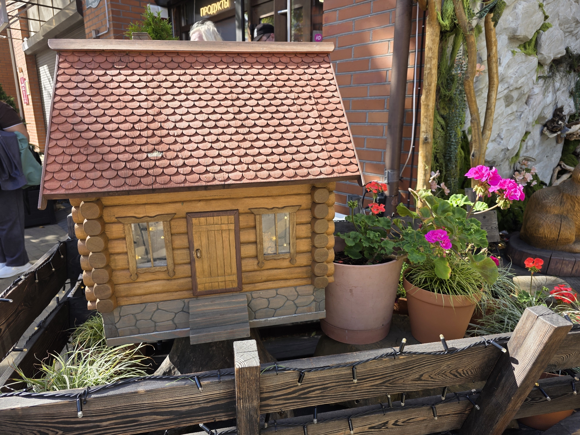 In Zelenogradsk, Russia, a miniature wooden log cabin, adorned with shingle-style roofing and stone-like detailing at its base, stands as a prominent feature. A wooden door and two small windows with decorative frames are visible on the cabin's facade. The cabin is surrounded by potted plants, including a large planter holding bright pink geraniums, and several smaller red flowers. The scene is framed by a rustic wooden fence, entwined with a string of small, white lights. In the background, brick building facades and a stone-like rock formation with moss and branches can be observed, suggesting an outdoor display or garden setting. Two individuals are partially visible in the upper left of the image, one wearing dark clothing and the other with blonde hair. The lighting suggests a bright, sunny day.