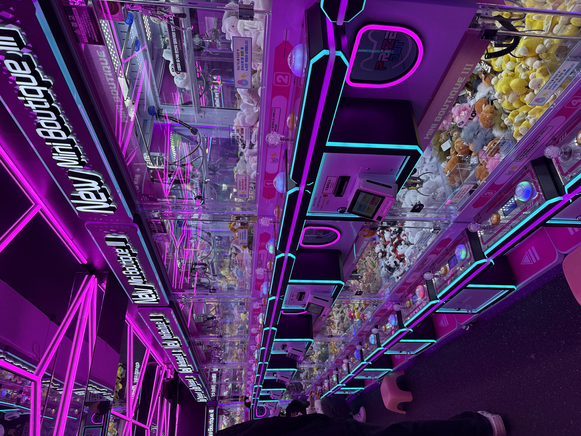 The image captures the interior of a "New Mini Boutique III" claw machine arcade in Seoul, South Korea. Rows of uniformly designed claw machines extend into the background, illuminated predominantly by magenta and cyan LED strip lighting. The ceiling features geometric patterns formed by magenta neon tubes. Each claw machine is a transparent enclosure containing various plush toys, keychains, and small collectible items. Control panels on each machine include a joystick, illuminated push buttons, a card reader, and a digital display. Korean text provides operational instructions and pricing, such as "1,000원 2회" (1,000 won for 2 plays). A "PRIZE OUT" chute is visible on the right side of some machines. Two individuals are partially visible: one person stands in the left foreground wearing dark pants and white sneakers, while another's hands are operating a machine's controls further back on the left. Pink plastic stools are positioned intermittently along the row of machines. The floor is dark and textured.