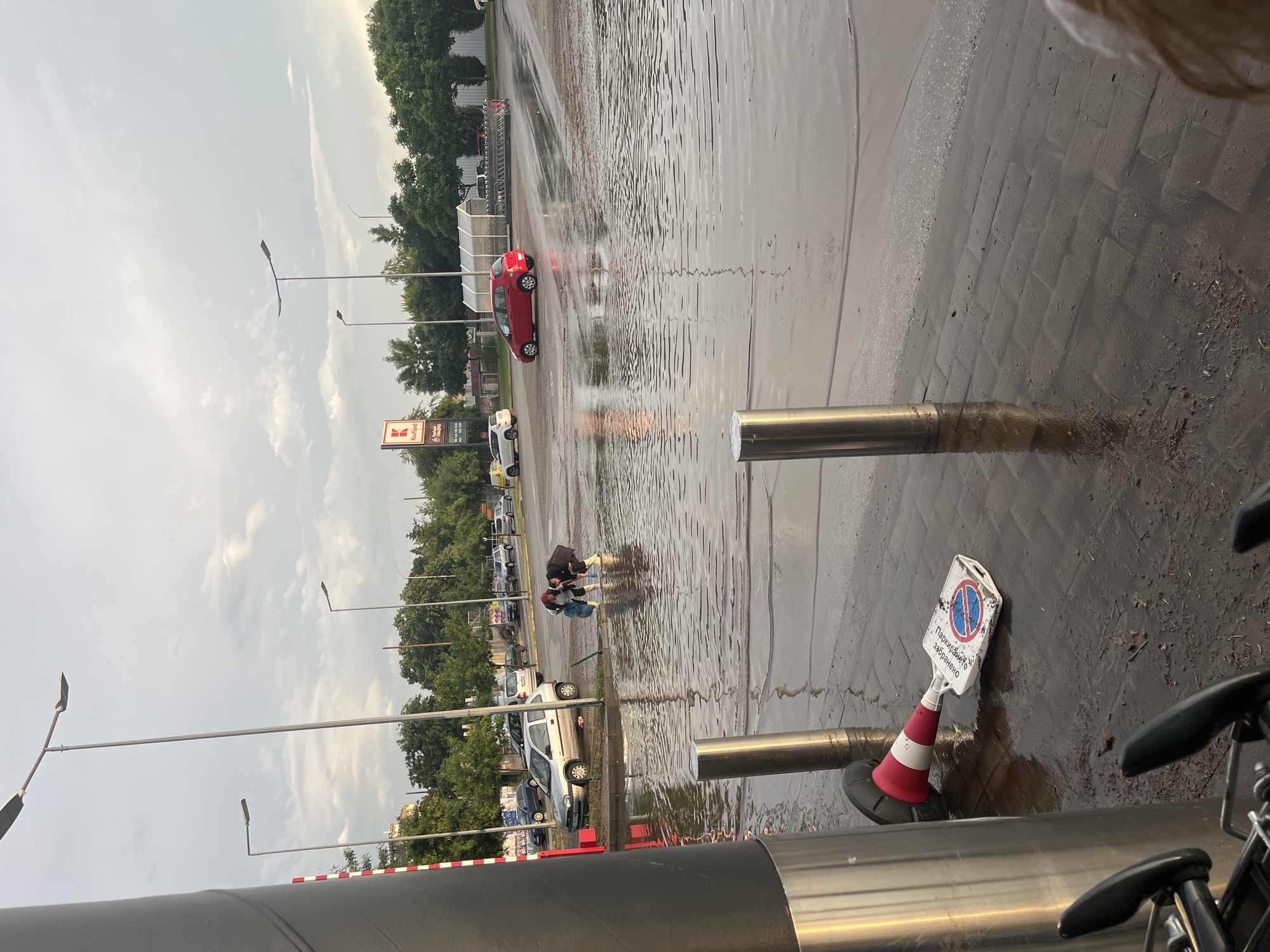 This image captures a flooded outdoor paved area, likely a supermarket parking lot, under an overcast sky. The perspective is from a slightly elevated position, looking across a wide expanse of standing water.
In the mid-ground, a red car is parked, partially submerged in the deep water, with visible ripples across the surface. Further back, several other cars are parked, also standing in the floodwaters. Two individuals are seen wading through the water in the middle of the scene, navigating the flooded conditions. They are dressed in dark clothing, and the water reaches at least their ankles, possibly higher.
To the left, a large red and white sign prominently displays the "Kaufland" logo, confirming the retail location. Beyond the parking area, a line of green trees and some distant buildings are visible. Multiple tall street light poles are scattered throughout the scene.
In the immediate foreground, to the right, a red and white traffic cone with an attached sign is partially submerged in muddy water. The sign features a red circle with a diagonal line, the universal symbol for "no parking." Below this symbol, text in Cyrillic script clearly reads "Паркирането забранено" (Parking forbidden). Two shiny metal bollards or posts are also visible in the foreground, reflecting the water's surface. The ground directly around these posts appears to be a mix of wet paved bricks and mud, contrasting with the smoother, flooded asphalt of the main parking area.
The sky is a uniform light grey, indicative of heavy cloud cover, strongly suggesting recent or ongoing heavy rainfall that caused the extensive flooding. The scene portrays the immediate aftermath of a significant downpour, disrupting activity in this commercial setting in Blagoevgrad, Bulgaria.