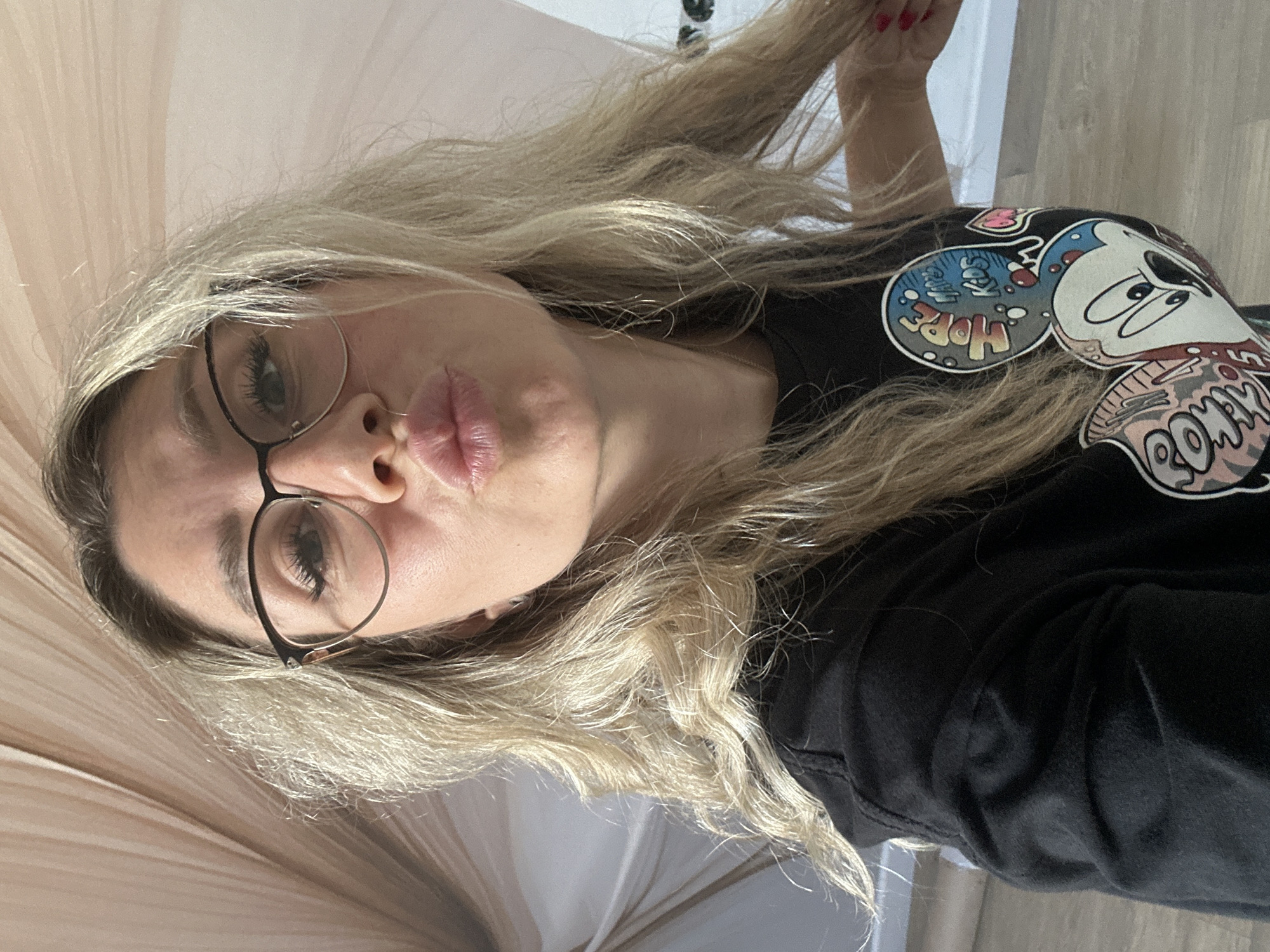 A woman with blonde hair and glasses, wearing a black t-shirt with a Mickey Mouse graphic, puckers her lips. The background appears to be indoors, with a light-colored fabric canopy overhead and a wooden floor. There is no discernible information to place this image in Novoaltaysk, Russia.