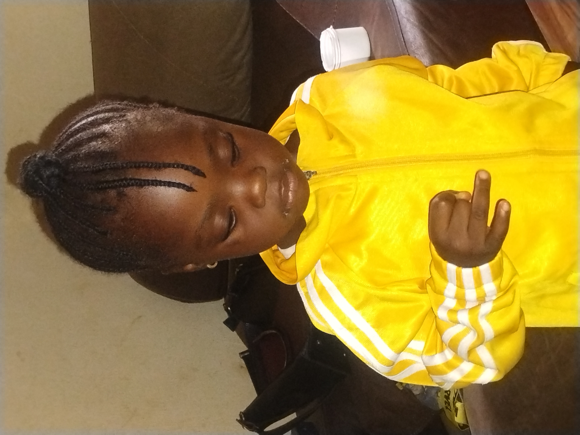 A child with braids, wearing a yellow tracksuit with white stripes on the sleeves, is lying down. The child's eyes are closed. One hand is visible, with the middle finger extended. A white cup is on the surface near the child. The background includes a brown leather-like surface and a beige wall. No specific information is provided to confirm the location in Jos, Nigeria.