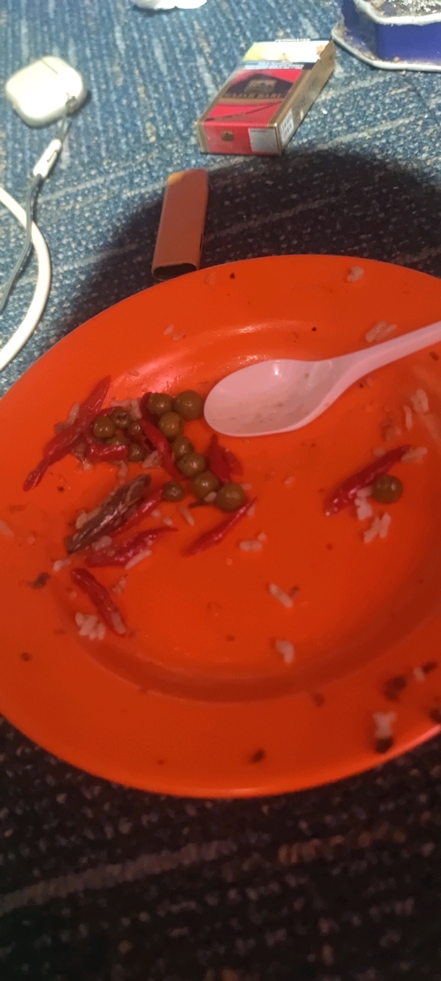 An orange plastic plate containing remnants of rice, green peas, and sliced red chilies sits on a dark, textured surface, possibly a carpet or rug. A white plastic spoon rests on the plate. To the left of the plate, a white object, possibly an earbud case, is connected to a white cable. In the upper right quadrant of the image, a pack of cigarettes and a lighter are visible. The overall scene suggests a casual setting, possibly a room, given the presence of a carpet-like surface and personal items. The specific location in Gambarsari, Indonesia, is not discernible from the visual elements of the image.