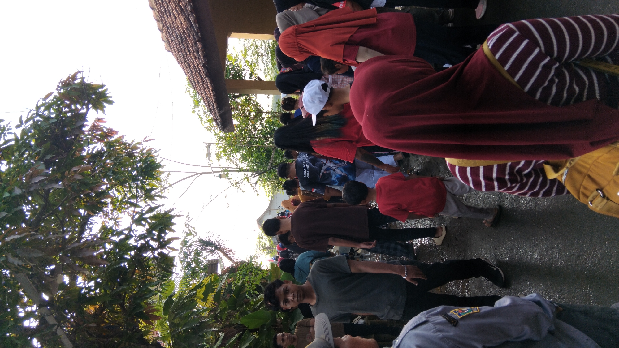 A group of people are gathered outdoors in what appears to be a street or pathway in the city of Posong, Indonesia. The ground is wet, suggesting recent rain. Trees with green and some brown leaves overhang the scene, and a building with a tiled roof is visible in the background. In the foreground, a young man with short dark hair is looking towards the camera, wearing a grey t-shirt. To his right, a person in a red t-shirt is walking away. Further into the crowd, individuals are seen wearing various clothing, including headscarves and t-shirts with graphic prints. The overall atmosphere suggests a gathering or event, possibly a procession or a public assembly.