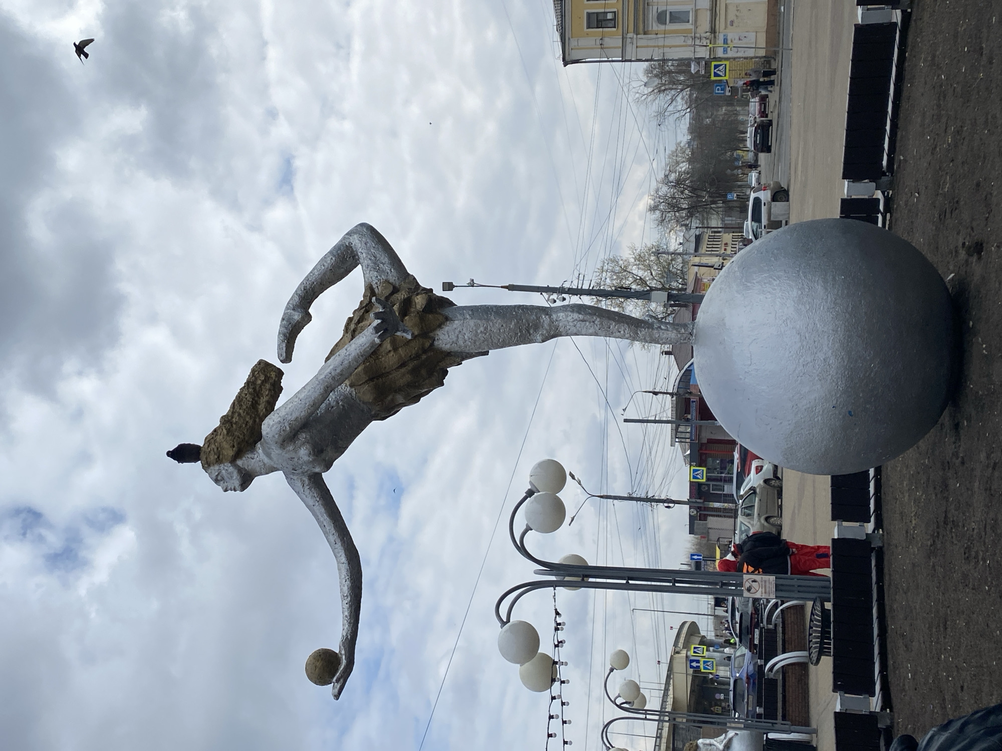 An outdoor photograph, taken in Nizhny Novgorod, Russia, captures a striking metallic statue under an overcast sky. The central subject is a large, silvery-grey, stylized female figure depicted in a dynamic pose, as if balancing or in motion. Her hair is notably textured, resembling rocks or rough earth. She extends her left arm, holding a small, rough, light-brown sphere. One foot of the statue appears to rest on a much larger, smooth, silvery sphere positioned on the ground to her right. A dark bird is perched on the statue's head, and another small bird flies in the cloudy sky above.
In the midground and background, an urban environment is visible. To the right of the large sphere, there's a paved area with black public benches, leading to a street occupied by several cars. A person wearing a bright orange vest and red pants is visible near the street. Traffic signs, including those for pedestrian crossings and parking ("P"), are discernible. The street is lined with older, light-colored (yellow/cream) buildings. To the left of the statue, modern streetlights with multiple spherical white lamps curve upwards into the frame. The ground in the immediate foreground consists of dirt or rough terrain. The scene is illuminated by the soft, diffuse light of an overcast day, with a sky filled with grey and white clouds.
