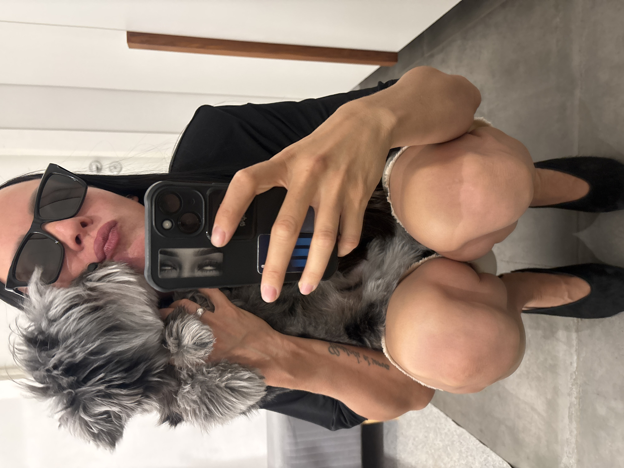 A woman is taking a selfie in what appears to be a bathroom. She is wearing dark sunglasses, a black top, and light-colored shorts or underwear. She's squatting or sitting low to the ground, hugging a gray-haired dog, a Schnauzer, and holding a smartphone. The phone has a dark case and what appears to be a photo of a person's eyes as a decoration. The background shows a white wall with a wooden strip, a gray tiled floor, and possibly a portion of a gray sofa or bed. The location and city are not identifiable.