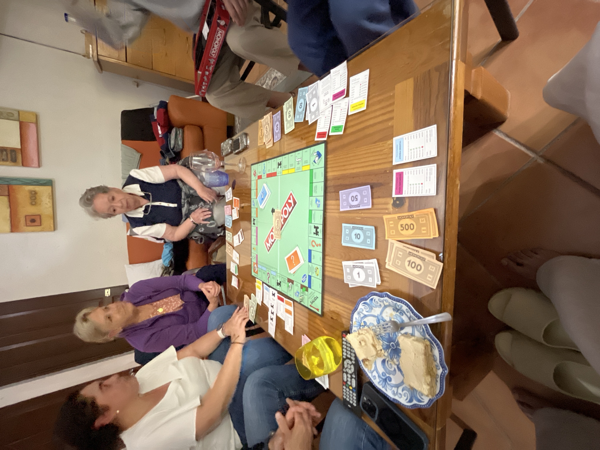 Four people are seated around a wooden table playing Monopoly. The Monopoly game board is on the table, with money and title deed cards scattered around it. One person, an older woman wearing a dark blue and white vest, is seated towards the back left of the table. Another woman, with blonde hair and wearing a purple cardigan, is seated to the left of the table. To the front left of the table sits a woman in a white shirt. There is a plate with a piece of cake and a fork, a yellow cup, a remote control, and a phone on the table. Also on the table is Monopoly money in the denominations of 1, 10, 50, 100, and 500. A fifth person can be seen sitting across from the older woman wearing a red backpack. The setting appears to be inside a home, with tiled floors and artwork on the wall. Based on the language of the Monopoly game, this image was likely taken in Puebla, Mexico.