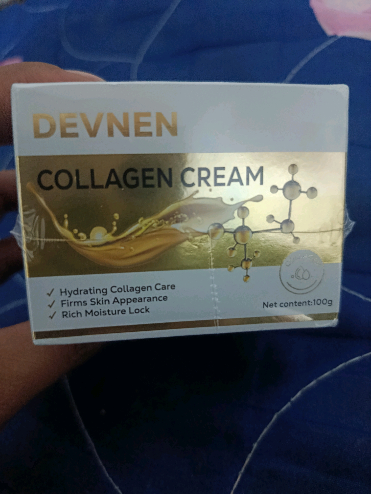 The image displays the front of a box for "DEVNNEN COLLAGEN CREAM." The box has a white background with gold lettering and accents. The text "DEVNNEN" is at the top in gold. Below that, "COLLAGEN CREAM" is also in gold lettering. There is an illustration of golden liquid splashing and a molecular structure. To the left of the illustration, there are three bullet points with checkmarks, listing "Hydrating Collagen Care," "Firms Skin Appearance," and "Rich Moisture Lock." In the bottom right corner, it states "Net content: 100g." A person's hand is visible on the left side of the image, holding or interacting with the box. The background appears to be a blue, textured fabric. There is no information about the location in Tambun Magih, Indonesia within the image itself.