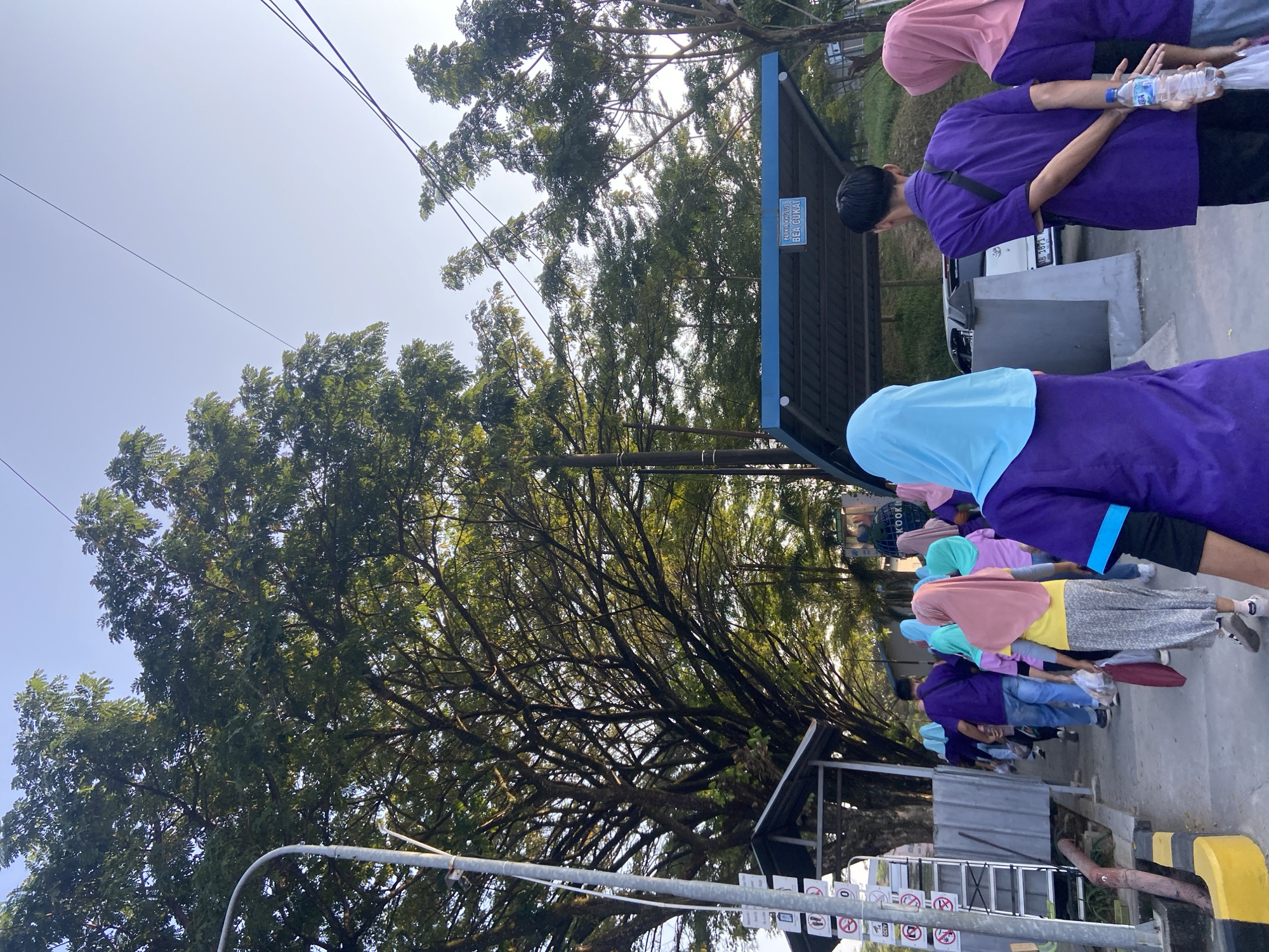A group of people, predominantly women wearing hijabs in various colors (pink, turquoise, and yellow) over purple shirts, walk along a paved path outdoors. They are accompanied by at least two men also in purple shirts. The path is bordered by a low concrete wall and a metal railing. To the left, a tall lamp post stands next to a building with a gray corrugated metal facade and a small covered area. A sign with multiple pictograms is affixed to the lamp post. Behind the people, a blue-roofed shelter with the text "PARKIRKHUSUS BEA CUKAI" is visible. A white car is parked under the shelter. The environment is characterized by lush green trees and foliage. The scene appears to be set in the city of Tanjungrejo, Indonesia, based on the signage. The lighting suggests it is daytime, possibly late afternoon due to the angle of the sunlight.
