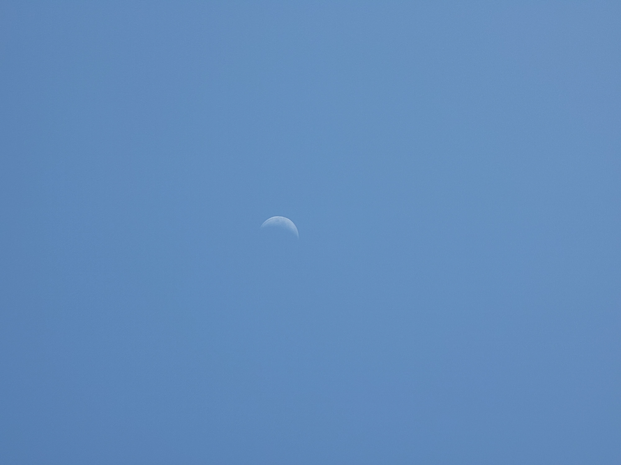A crescent moon is visible in a clear blue sky. The image was taken in Quarteira, Portugal. There are no people, objects, or actions depicted in the scene. The environment is simply the sky.