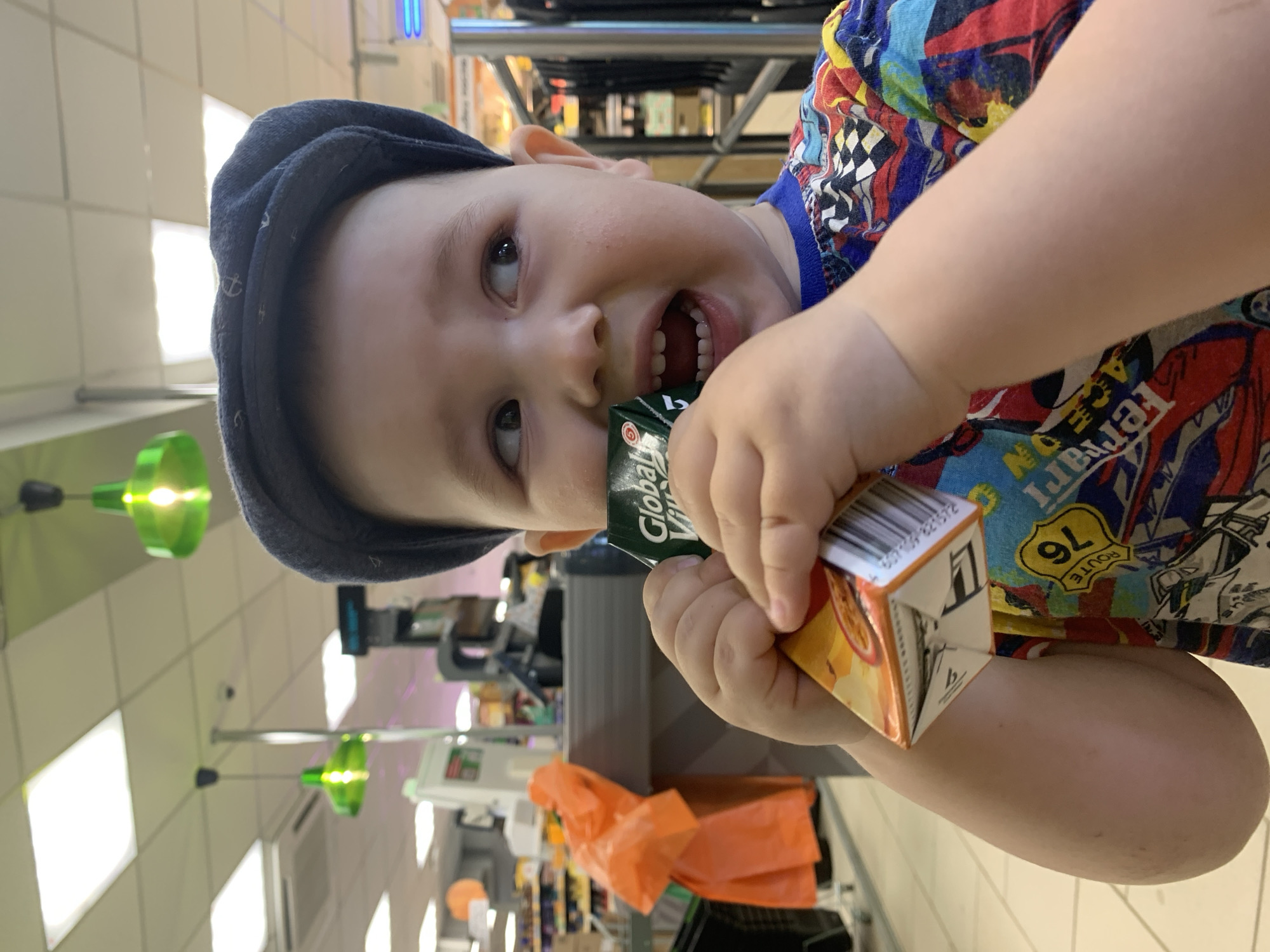 A child in a baseball cap and patterned shirt is inside a retail environment. He is holding a carton of orange juice in his right hand and a smaller box, possibly another beverage, in his left hand, holding it up to his mouth as if about to bite it. The location appears to be a supermarket aisle, with rows of shelves and other products visible in the background. The lighting is bright and the aisle appears to be relatively empty. The overhead lights are green. An orange plastic bag is present near the foreground.