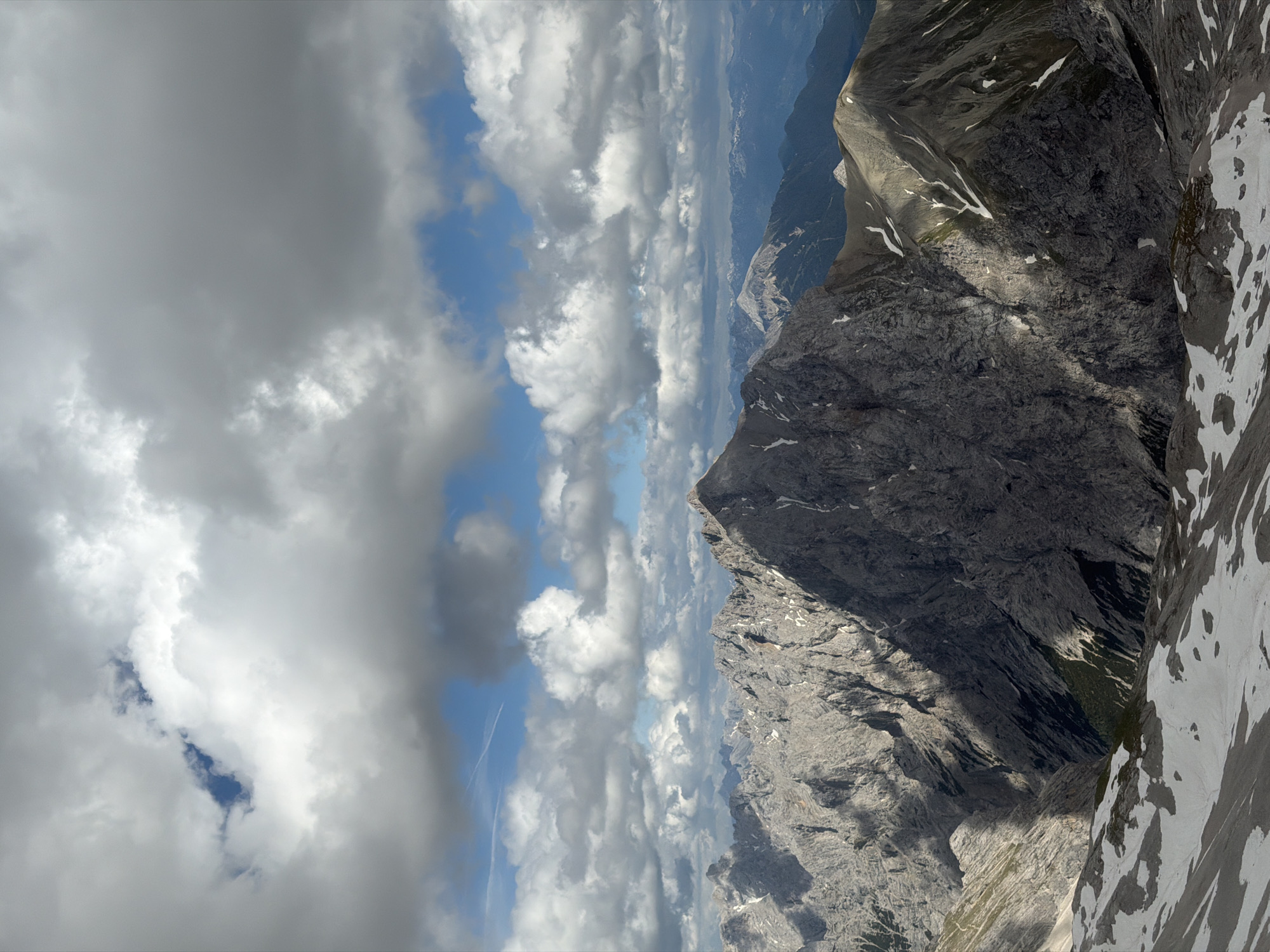 The image depicts a dramatic mountain landscape under a partly cloudy sky, likely taken from a high vantage point in or near Garmisch-Partenkirchen, Germany. The foreground is characterized by rugged, rocky terrain interspersed with patches of snow, suggesting a high-altitude alpine environment.
Dominating the mid-ground is a large, imposing mountain peak with steep, gray, rocky slopes. Sunlight casts dramatic shadows across its surface, highlighting the textures and crevices of the rock. To the right of this central peak, another mountain ridge is visible, also with rocky slopes and streaks of snow. This ridge slopes downwards towards the background.
In the background, a series of layered mountain ranges recede into the distance. The furthest mountains appear softer and bluer, indicating atmospheric perspective. The sky above is a mix of bright blue sky and large, voluminous white and gray clouds. Some of these clouds are illuminated by the sun, while others are darker, suggesting they are thicker or casting shadows. A faint contrail from an aircraft can be seen in the upper left portion of the sky.
The overall impression is one of grand natural scenery, likely a view from a mountain summit or a high ridge in the Bavarian Alps. Given the characteristic scenery, this photograph could be from the Zugspitze area or a similar prominent peak accessible from Garmisch-Partenkirchen. No people or man-made structures are visible in the scene. The environment is wild and natural, dominated by rock, snow, and sky.