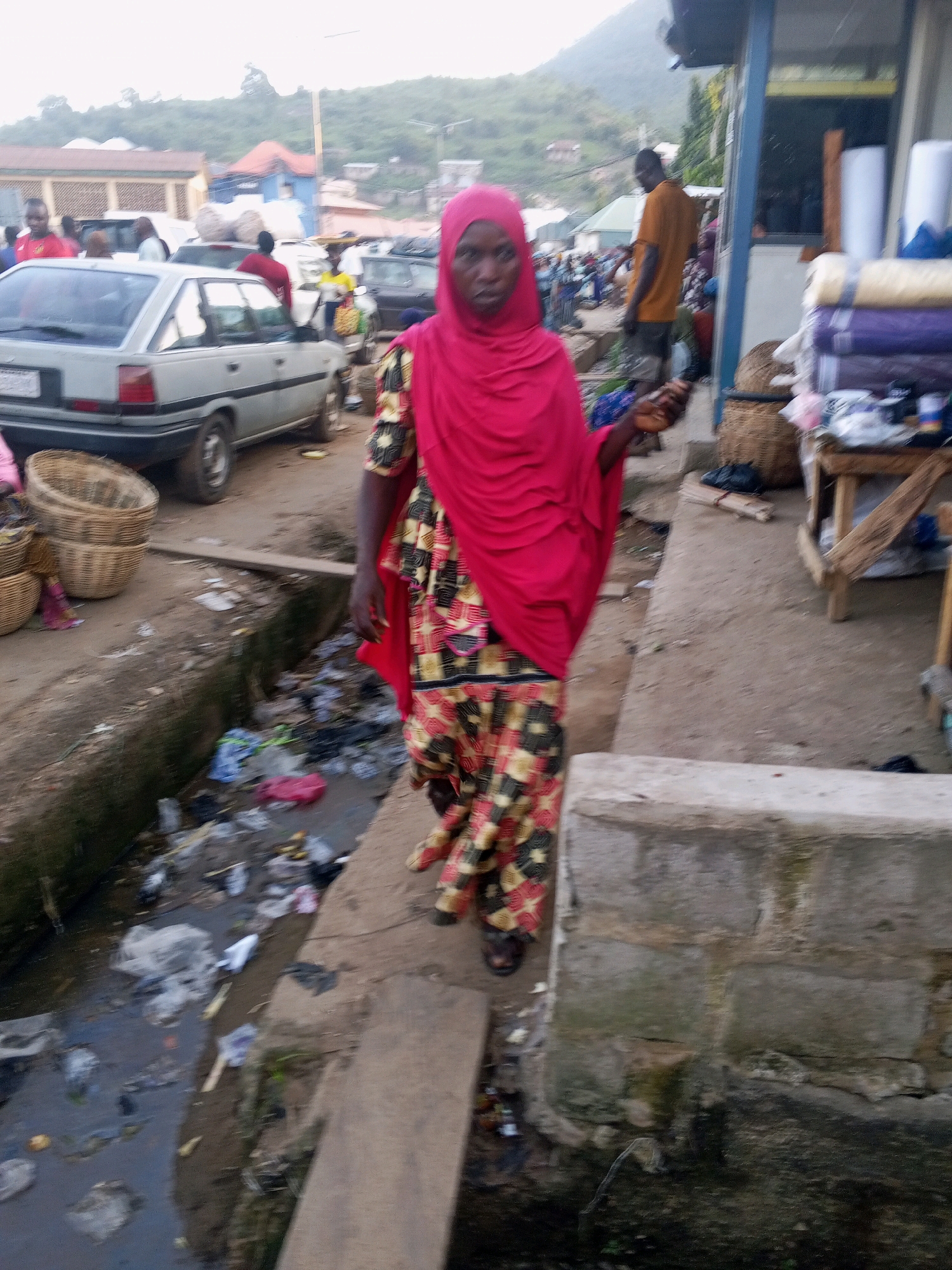 The image depicts a street scene in Lokoja, Nigeria, characterized by a busy market environment and visible signs of disrepair. A woman, wearing a bright pink hijab and a colorful patterned dress, is the central figure, walking towards the camera. She has dark skin and appears to be looking directly at the viewer.
In the background, various elements suggest a typical urban market setting. Several cars are parked or moving on the unpaved road, including a silver sedan. People are scattered throughout the scene, some appearing to be vendors or shoppers, although their faces are not clearly discernible. Buildings line the street, some with shops displaying goods. To the right, a stall is set up with rolls of fabric or textiles.
The environment shows signs of neglect. A narrow, open drain runs along the side of the road, filled with stagnant water and considerable amounts of garbage, including plastic bags, wrappers, and other debris. The ground is uneven and dusty. In the foreground, stacked woven baskets are visible, as is a wooden plank that appears to serve as a makeshift walkway over the drain. Hills and vegetation are visible in the distance, characteristic of the landscape around Lokoja. The overall atmosphere is one of a bustling, yet somewhat unsanitary, urban marketplace.