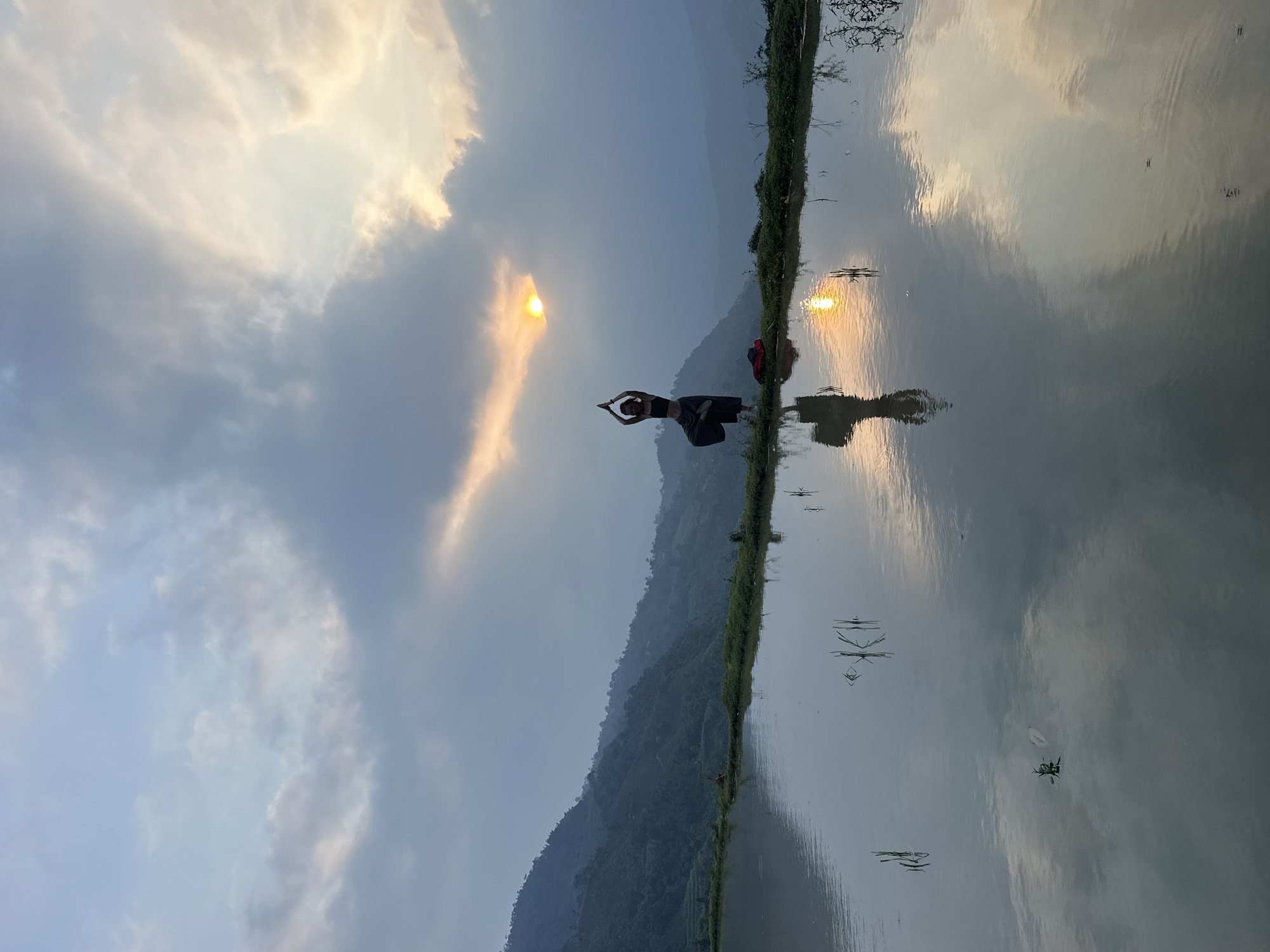 The image captures a serene outdoor scene at either sunrise or sunset in a mountainous region, likely Nậm Dịch Commune, Vietnam. A person, appearing to be a woman, is centered in the frame, standing on a narrow grassy bank that separates a calm body of water from a backdrop of lush, rolling hills. She is performing a balancing yoga pose, possibly Tree Pose, with her arms raised above her head, palms pressed together.
The sun, a bright golden disc, is low in the sky, partly veiled by soft clouds, casting a warm, ethereal glow. Its intense light is dramatically reflected on the still surface of the water, mirroring the sun, clouds, and the distant mountains. The person's reflection is also visible in the water. The sky displays a gradient of soft blues, grays, and golden hues. Small ripples and a few dark specks, possibly water insects or debris, are visible on the water's surface. A red bag or piece of clothing lies on the bank near the person's feet. The overall ambiance is peaceful and contemplative, highlighting a connection with nature. No visible text is present in the image.