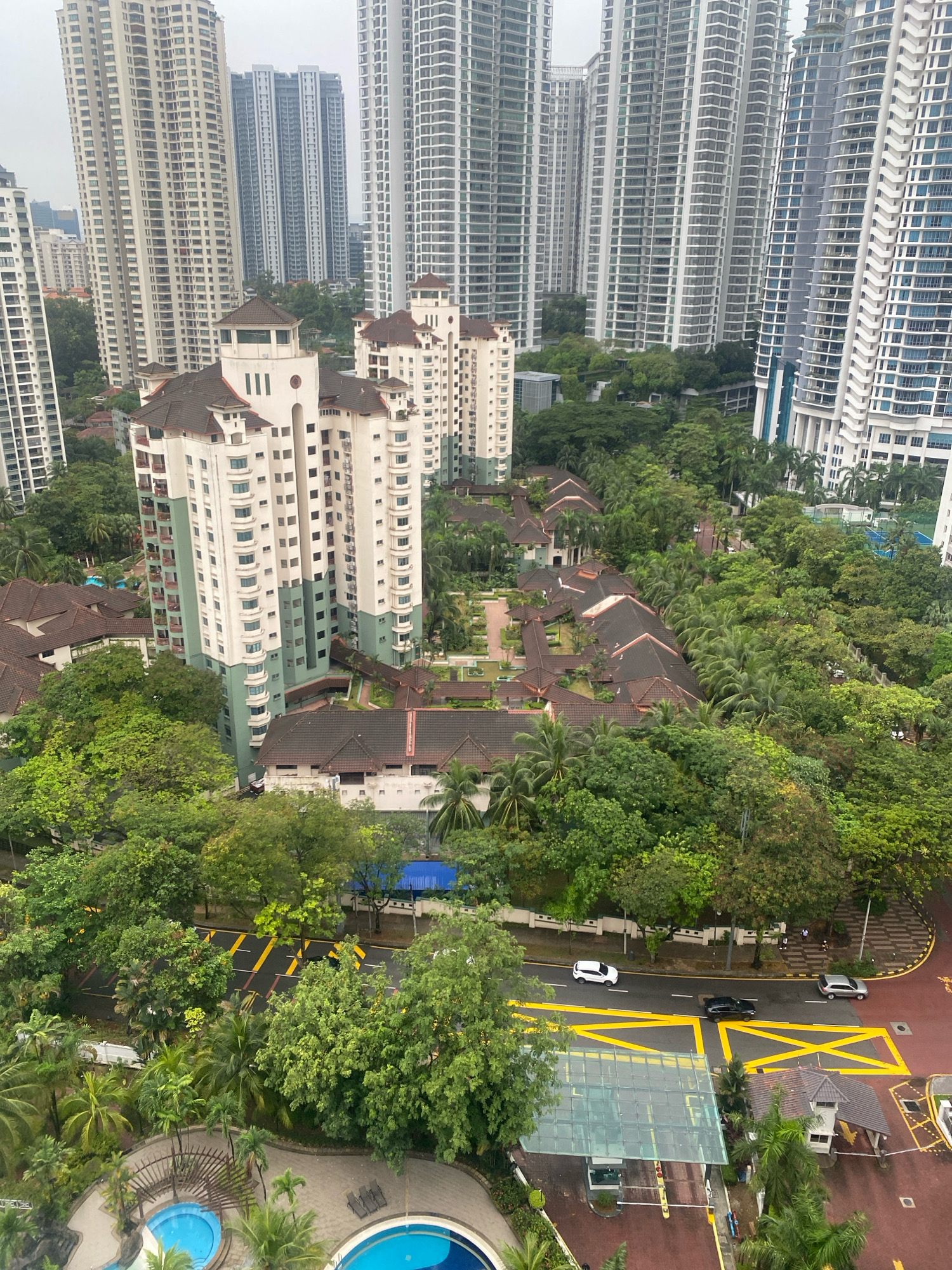 An aerial view captures a dense urban landscape in Kuala Lumpur, Malaysia, dominated by high-rise apartment buildings and lush greenery. The foreground features two resort-style swimming pools with blue water, surrounded by paved areas and palm trees. A short distance away, a complex of lower-rise residential buildings with dark tiled roofs is nestled amongst the trees.
Further back, a multi-lane road runs horizontally across the frame, with yellow road markings and several vehicles visible, including a white car, a black car, and a silver car. The road is lined by a white wall and a dense canopy of green trees, creating a stark contrast with the built environment.
In the background, towering skyscrapers with modern architectural designs fill the horizon, their numerous windows reflecting the overcast sky. The overall impression is one of a city that balances high-density living with significant green spaces. The scene appears to be captured on an overcast day, contributing to a soft, diffused light.