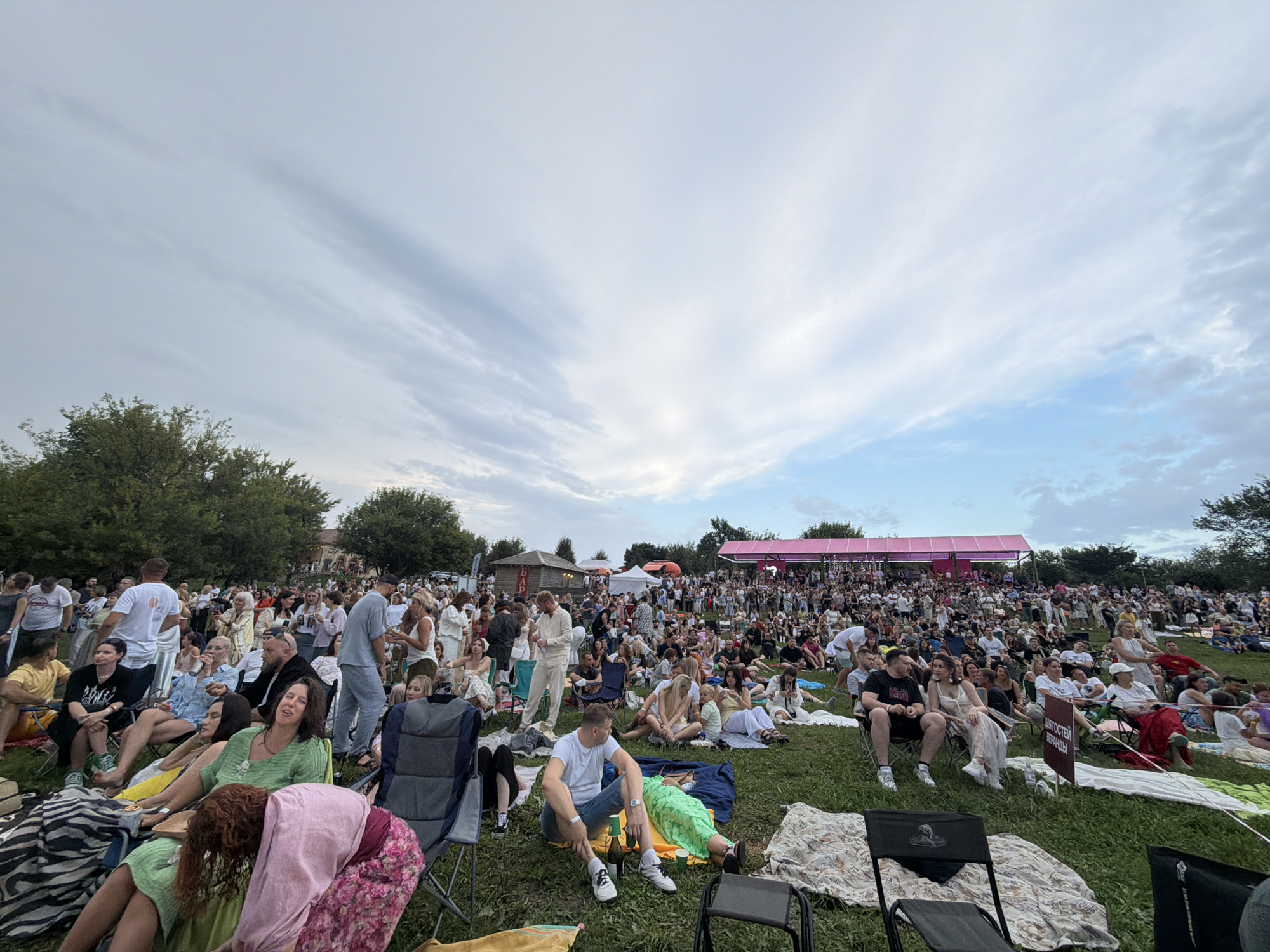 The image depicts an outdoor gathering of a large crowd of people sitting on a grassy slope. In the background, there is a large pink structure, possibly a stage or canopy, and several trees line the horizon. The sky is cloudy with patches of blue. The people are dressed in casual clothing and are seated on chairs, blankets, and the grass, suggesting a festival, concert, or similar event. The overall atmosphere appears relaxed and communal. The specific location within Skoryakovo, Russia, is not identifiable from the visual information alone.