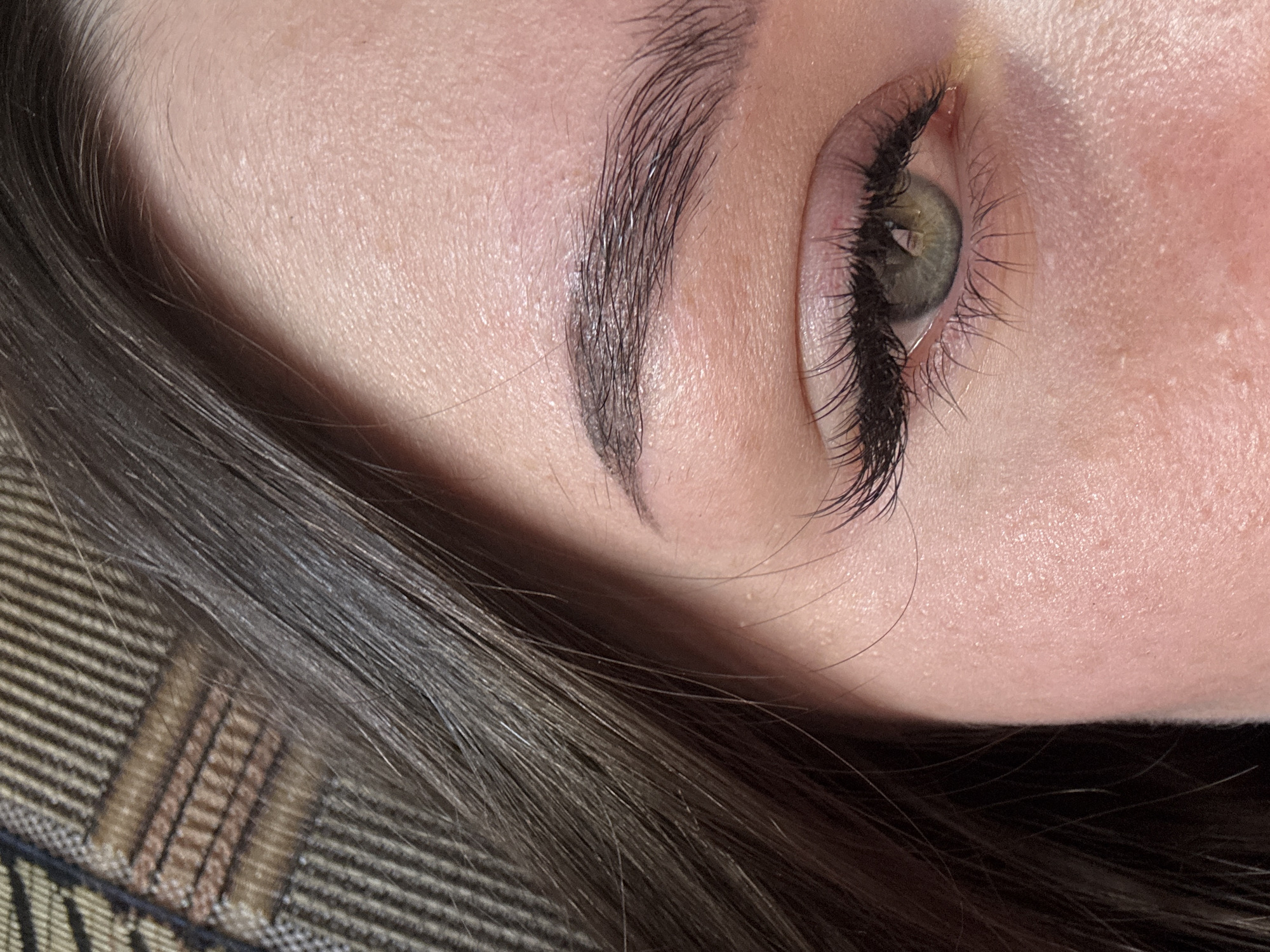 This is a close-up, angled photograph of a person's eye and eyebrow. The subject has fair skin with visible pores. Their eye is open, revealing a green iris. Long, dark eyelashes frame the eye, and a dark eyebrow arches above it. The person's dark hair is visible in the foreground, partially obscuring the lower part of the image. The background appears to be a striped fabric, possibly a blanket or upholstery. The lighting suggests an indoor setting, and the focus is sharp on the eye and eyebrow, with the hair slightly out of focus. There are no visible text elements in the image. The overall impression is one of a personal, intimate portrait focused on facial features. The crops highlight different areas of the face and hair, emphasizing the texture of the skin, the individual hairs of the eyebrow and eyelashes, and the weave of the fabric.