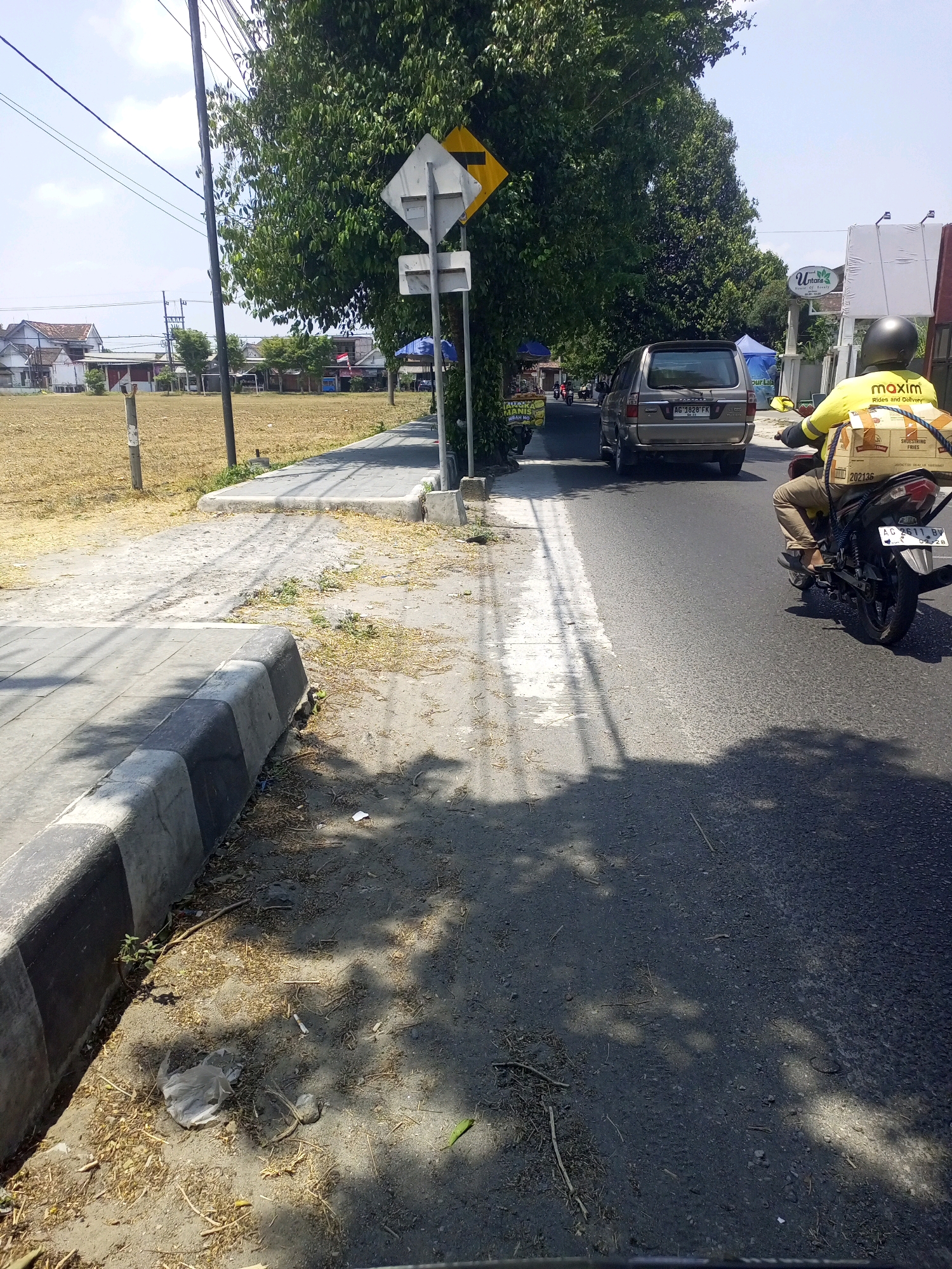 In Ngasem, Indonesia, a motorcycle courier wearing a yellow jacket with "maxim" branding and a helmet rides on a paved road. The courier is transporting a large, rectangular box strapped to the back of the motorcycle. Ahead of the courier, a silver SUV with license plate AG 1828 FK is also traveling on the road. The scene is bordered by a sidewalk with a grey and black curb on the left and a grassy area with scattered debris on the right. Trees with dense green foliage line the right side of the road, casting shadows on the asphalt. A traffic sign with a yellow diamond and a white diamond shape is visible near the trees. In the background, buildings and houses are visible, along with utility poles and electrical wires. The sky is clear and blue.