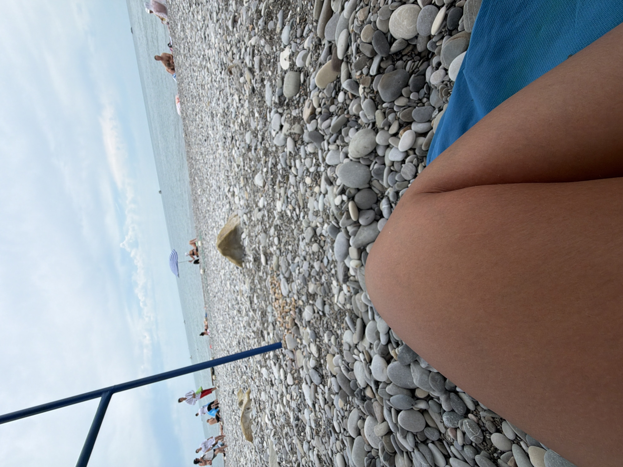 The image captures a view of a pebble beach in Sochi, Russia. In the foreground, a person's legs and thighs are visible, resting on a blue towel. The background features a wide expanse of gray and white pebbles leading to the sea. Several people are scattered across the beach, some sitting or lying down, while others are standing or walking. A blue umbrella provides shade for some beachgoers. The sea stretches out to the horizon under a cloudy sky. A dark blue pole, part of a beach structure, is visible on the left side of the frame.
