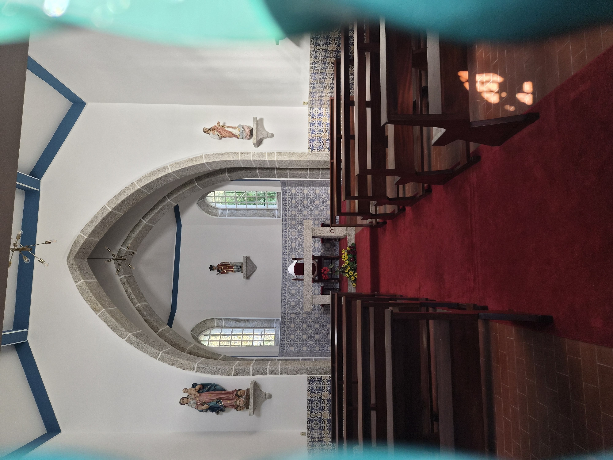 The image shows the interior of a church, likely the Igreja Matriz de Marinhas in Esposende, Portugal, based on architectural details. Rows of wooden pews are on either side of a red carpeted aisle. At the far end is an altar decorated with flowers, against a wall adorned with blue and white tiles. The walls are white with blue trim. Sculptural figures are mounted on the walls. Natural light filters through arched windows.