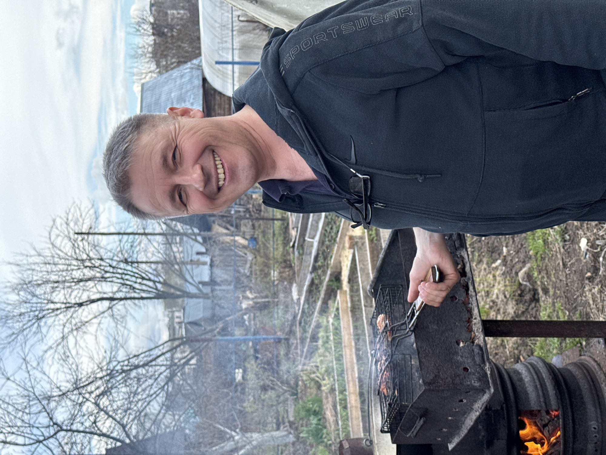 An outdoor, portrait-oriented image taken in Chaikovsky, Russia, captures a smiling man grilling food. The man, with short, graying hair, is positioned in the foreground, facing the camera with a wide, cheerful smile that shows his teeth. He is wearing a dark hooded jacket, visible text "SPORTSWEAR" and a Nike swoosh logo on the left sleeve, and glasses are tucked into the front of the jacket. Underneath, he wears a dark shirt.
He is actively engaged in grilling, holding a metal skewer or tong to tend to food (likely meat) cooking on a grate over a metal brazier. Flames and smoke rise from the hot coals within the grill.
The setting appears to be a rural or suburban backyard or garden area. The background features bare trees, suggesting late winter or early spring. Raised garden beds or similar structures are visible in the mid-ground. The sky is overcast, and the overall lighting indicates daytime. The man's joyful expression conveys a positive and relaxed atmosphere.