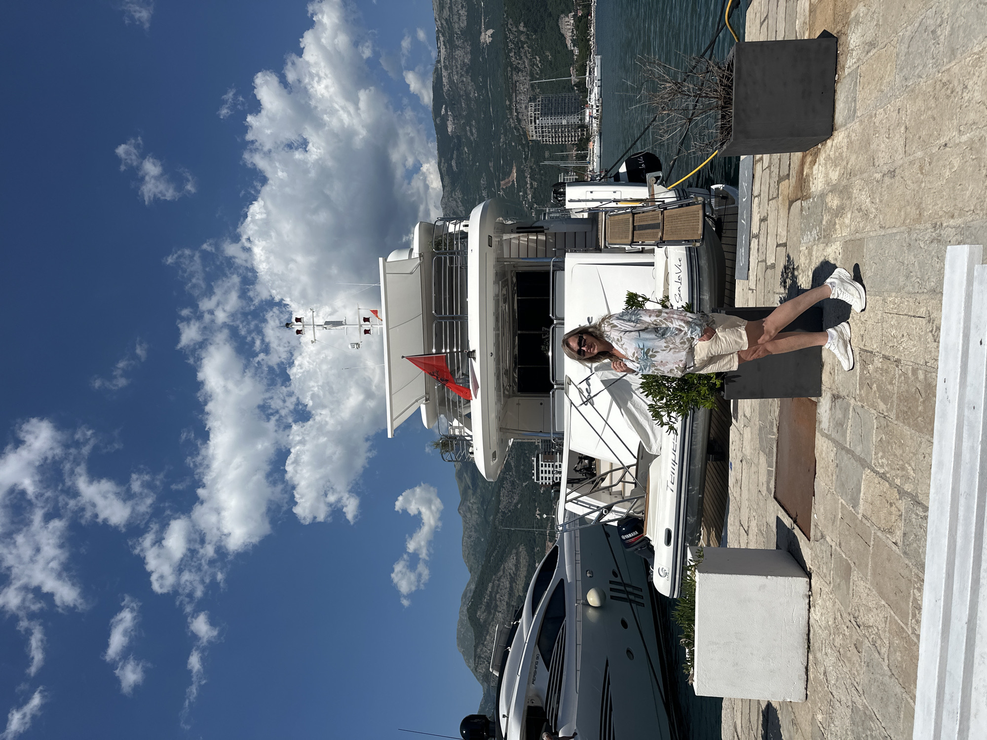 The image depicts a sunny outdoor scene, likely taken in Budva, Montenegro, given the presence of recognizable mountainous terrain and coastal setting.
A woman stands on a stone-paved area near a modern yacht. The yacht is mostly white with dark windows and is docked in a harbor, with other boats visible in the background. The yacht is labeled "PERISHING 90" on the lower-left, and the name "Tempest" and "Sea La Vie" are marked on the smaller white boats in the background. Mountains and buildings are partially visible in the background, suggesting a coastal city.
The woman is wearing a floral shirt, light-colored shorts, and white sneakers. She appears to be posing for the photo. Above, a bright blue sky is filled with scattered white clouds. The camera is tilted on its side, causing the vertical orientation of the image.