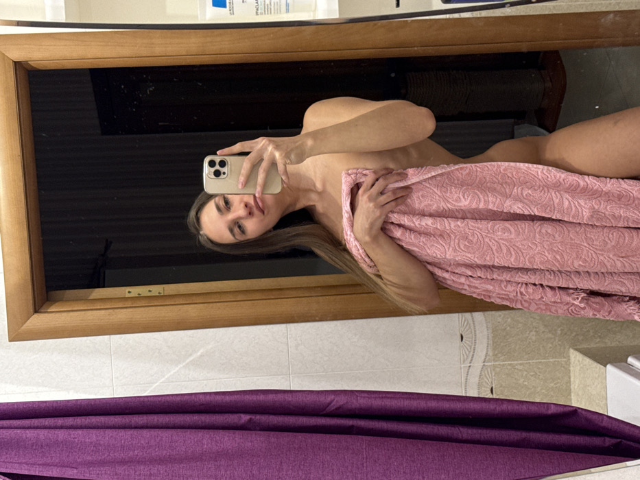 A mirrored image shows a woman holding a pink towel, concealing her body. She holds a smartphone to take a selfie. The background reveals a bathroom setting with light-colored tiles and a purple shower curtain below the mirror. Within the mirror's reflection, a shelf or ledge holds various products.