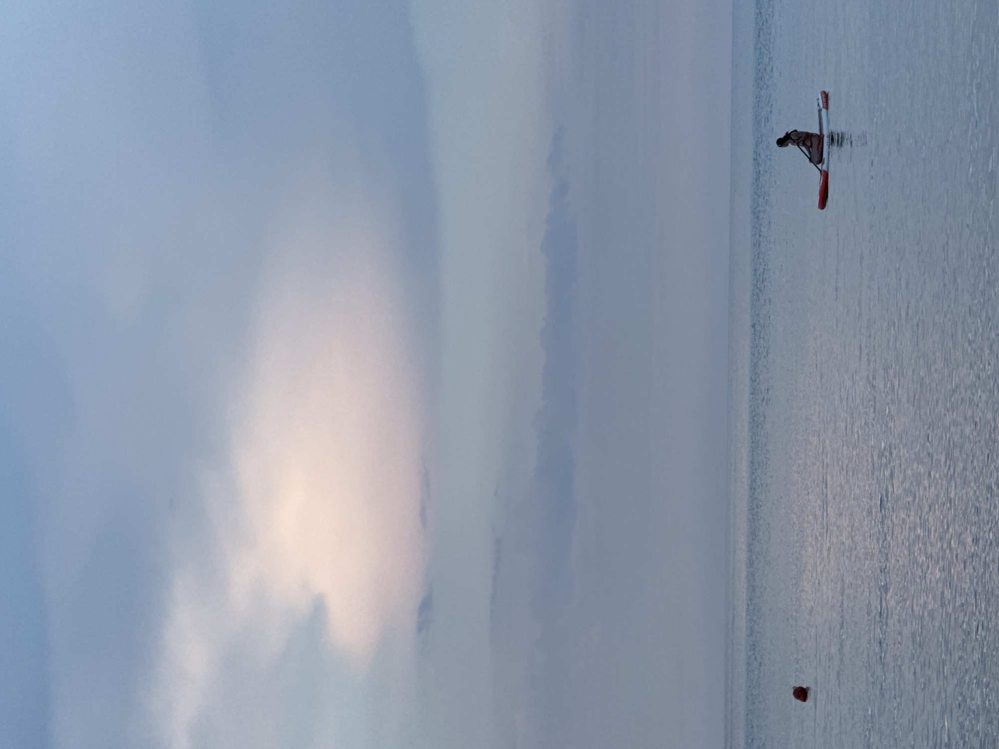 A lone figure is paddleboarding on a calm sea, with a red buoy visible to the left of the frame. The sky above is filled with soft, diffused clouds, hinting at either dawn or dusk. The water is a muted blue-grey, reflecting the colors of the sky. The paddleboarder, silhouetted against the water, is seated on the board and appears to be holding a paddle. The scene evokes a sense of solitude and tranquility. This image may depict a location in or near Partenit, Ukraine, a coastal town known for its scenic beauty.