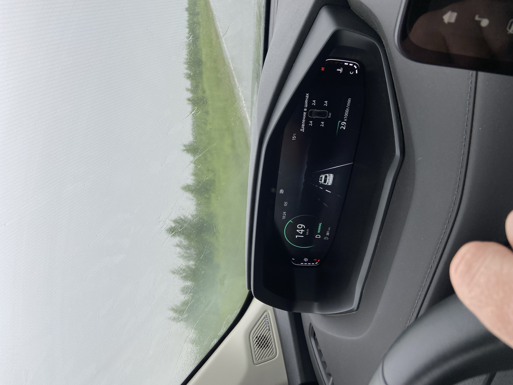 The image shows the interior of a car, specifically the dashboard and the view through the windshield, likely taken from the driver's perspective. The car is in motion, indicated by the speed of 149 km/h displayed on the digital dashboard. The weather appears to be overcast and rainy, with raindrops visible on the windshield. The dashboard displays various information including tire pressure (2.4 bar), engine RPM (2.9 x1000r/min), and the current gear (D for Drive, NORMAL mode). Outside the car, a green, grassy roadside with trees can be seen. The location is identified as Поварёнки, Russia based on the context. A hand is visible in the lower left corner, suggesting the driver is operating the vehicle. A portion of a smartphone or secondary display is visible in the lower right corner.