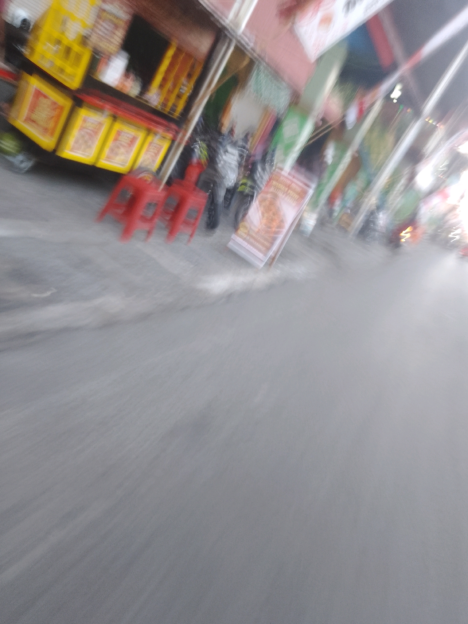 The image captures a street scene in Sedati Agung, Indonesia, characterized by motion blur, suggesting movement. In the foreground, a grey asphalt road is visible, with diagonal streaks indicating the direction of motion. A row of three red stools is present, also blurred. Behind the stools, a food stall is depicted with yellow and red elements, possibly advertising products. Several blurry figures, likely people and motorcycles, are present around the stall and further down the street. Banners and signs with various colors and designs are displayed, adding to the vibrant, albeit blurry, streetscape. The overall impression is of a bustling urban environment in motion.