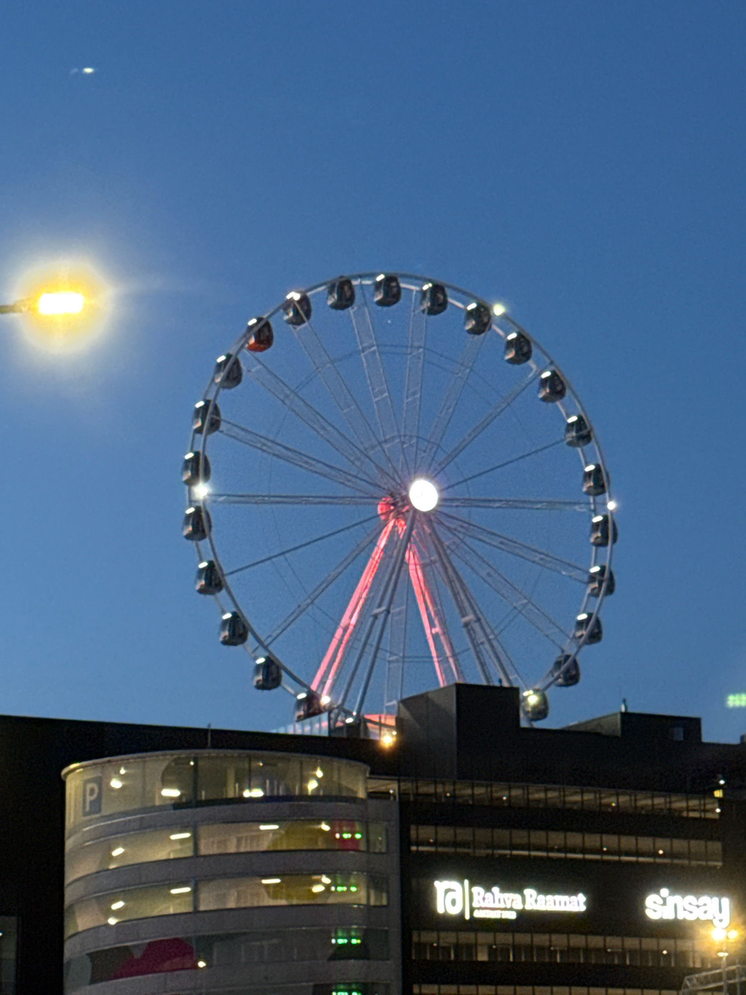 The image shows a ferris wheel illuminated with red and white lights against a dark blue sky. The ferris wheel is located in the city of Tallinn, Estonia, and is positioned above a modern building complex. The building has a curved section with a visible "P" sign, indicating parking, and multiple floors with illuminated windows. The building also features lit signage for "Raha Raamat" and "Sinsay". A street lamp with a bright yellow light is visible in the upper left corner of the image. The overall scene suggests an urban evening or nighttime setting.